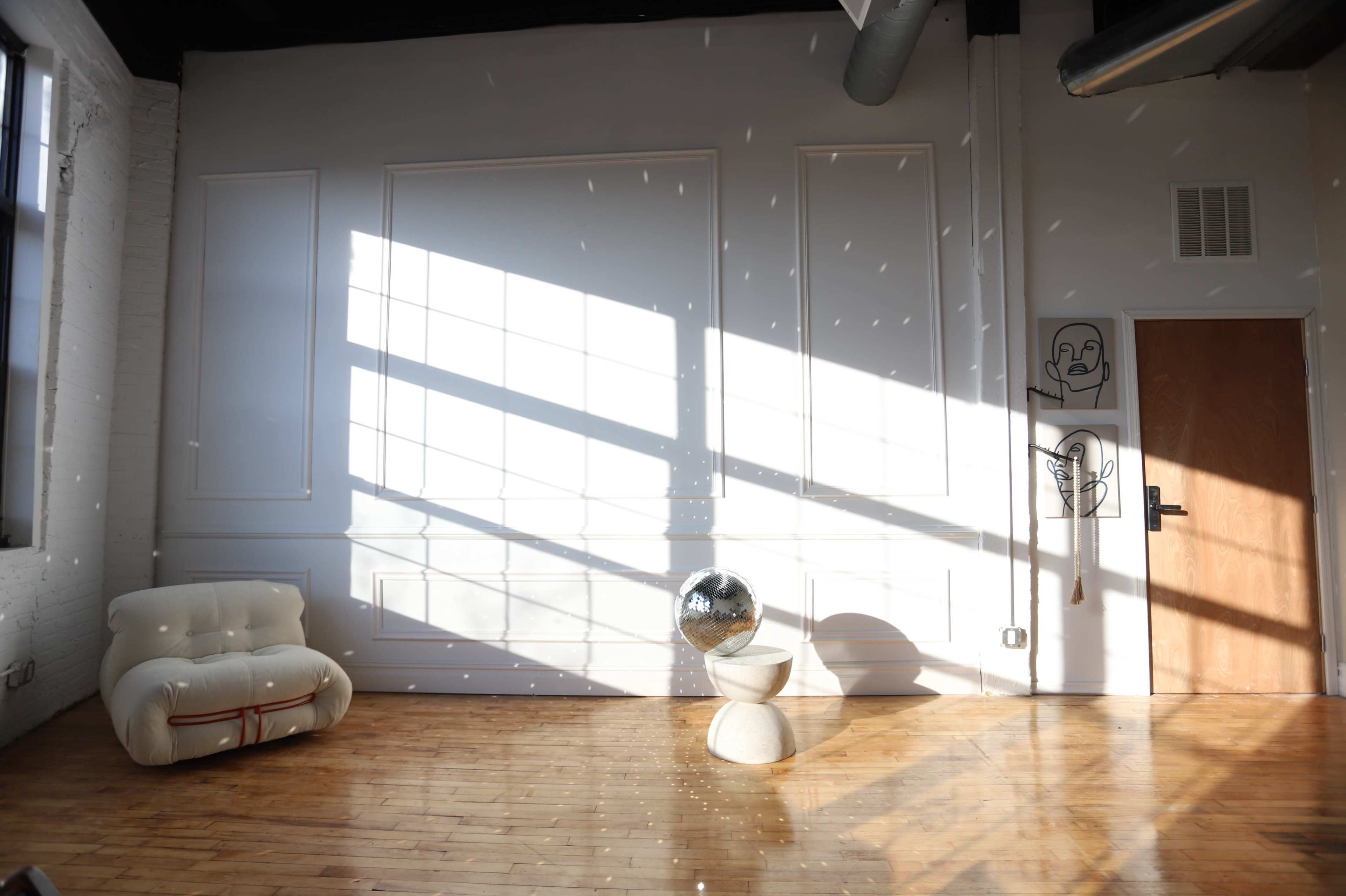 A sunlit room features a white curved sofa, a reflective disco ball on a pedestal, and shadows cast by large windows.