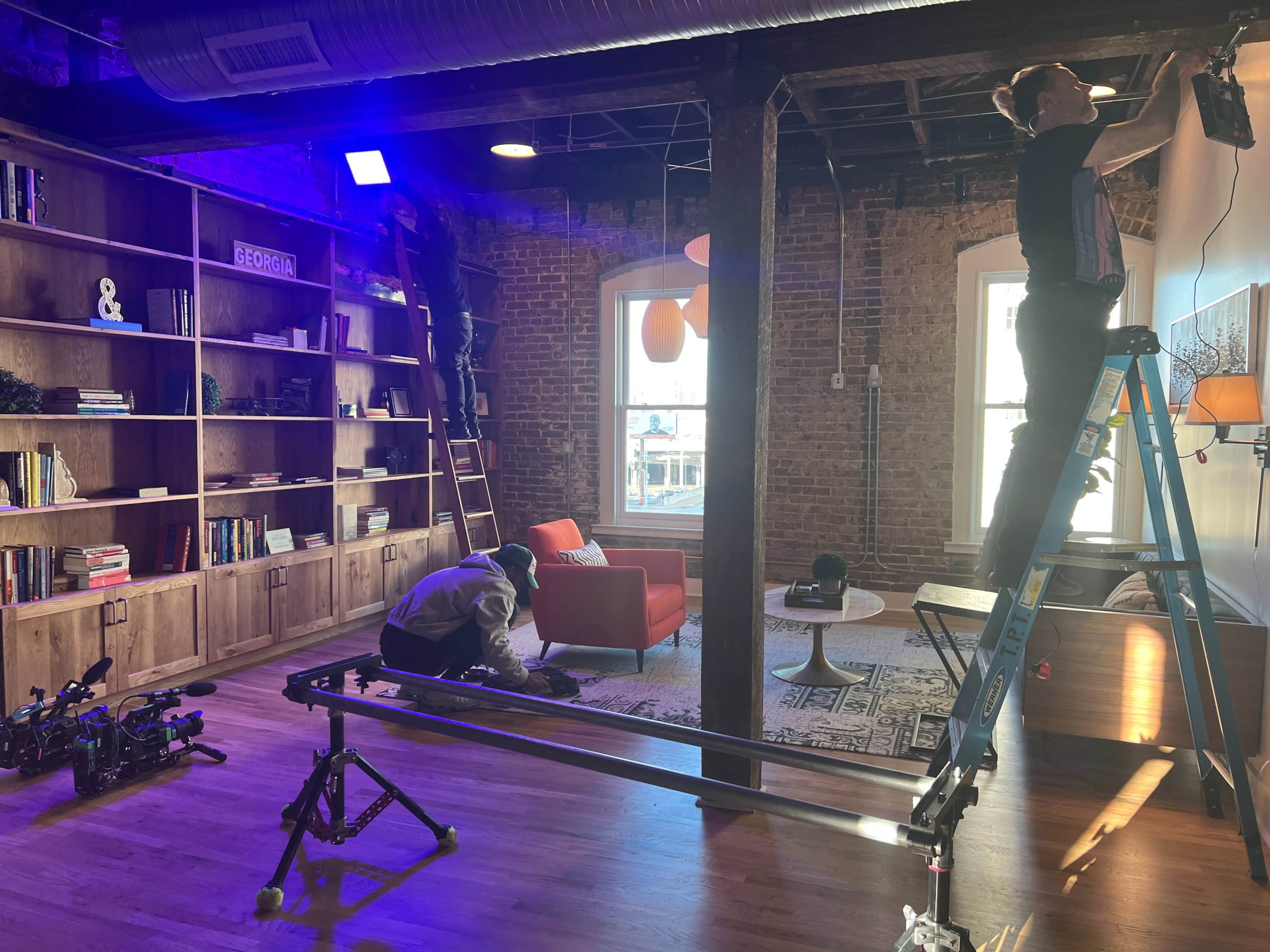 Two crew members are setting up a film scene in a brightly lit room with a large bookshelf, a red sofa, and a camera dolly on the floor.