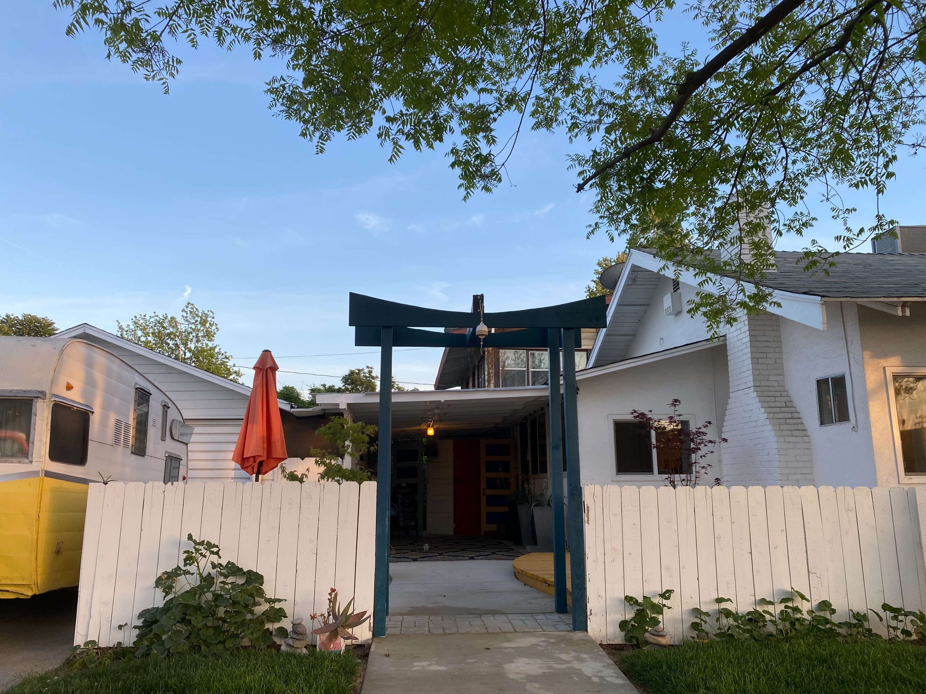 A white picket fence surrounds a house with a carport and a vintage camper trailer, featuring an orange umbrella on the patio.