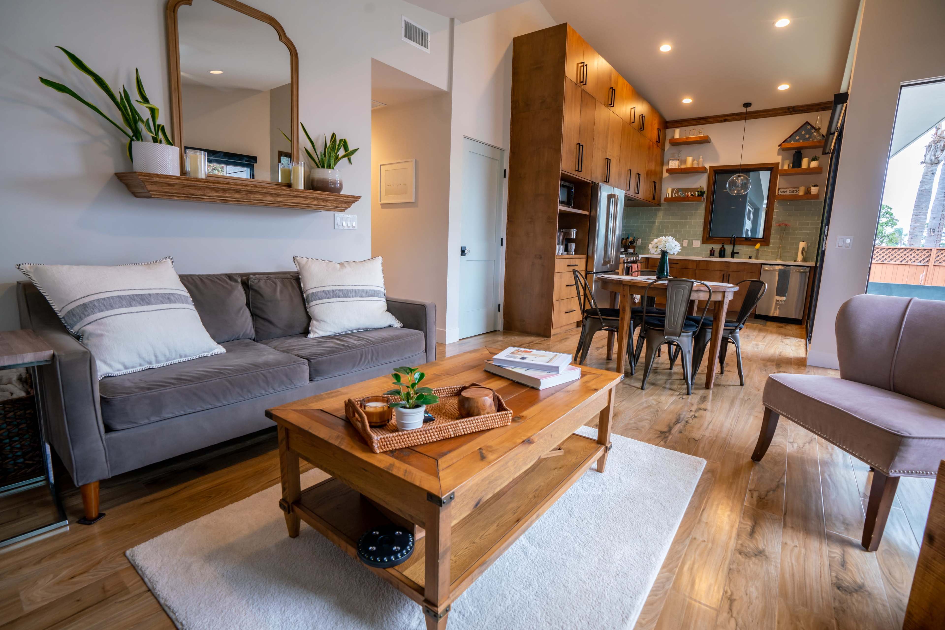 The image shows a cozy living space featuring a gray sofa, a wooden coffee table with decorative items, and a dining area with a modern kitchen in the background.