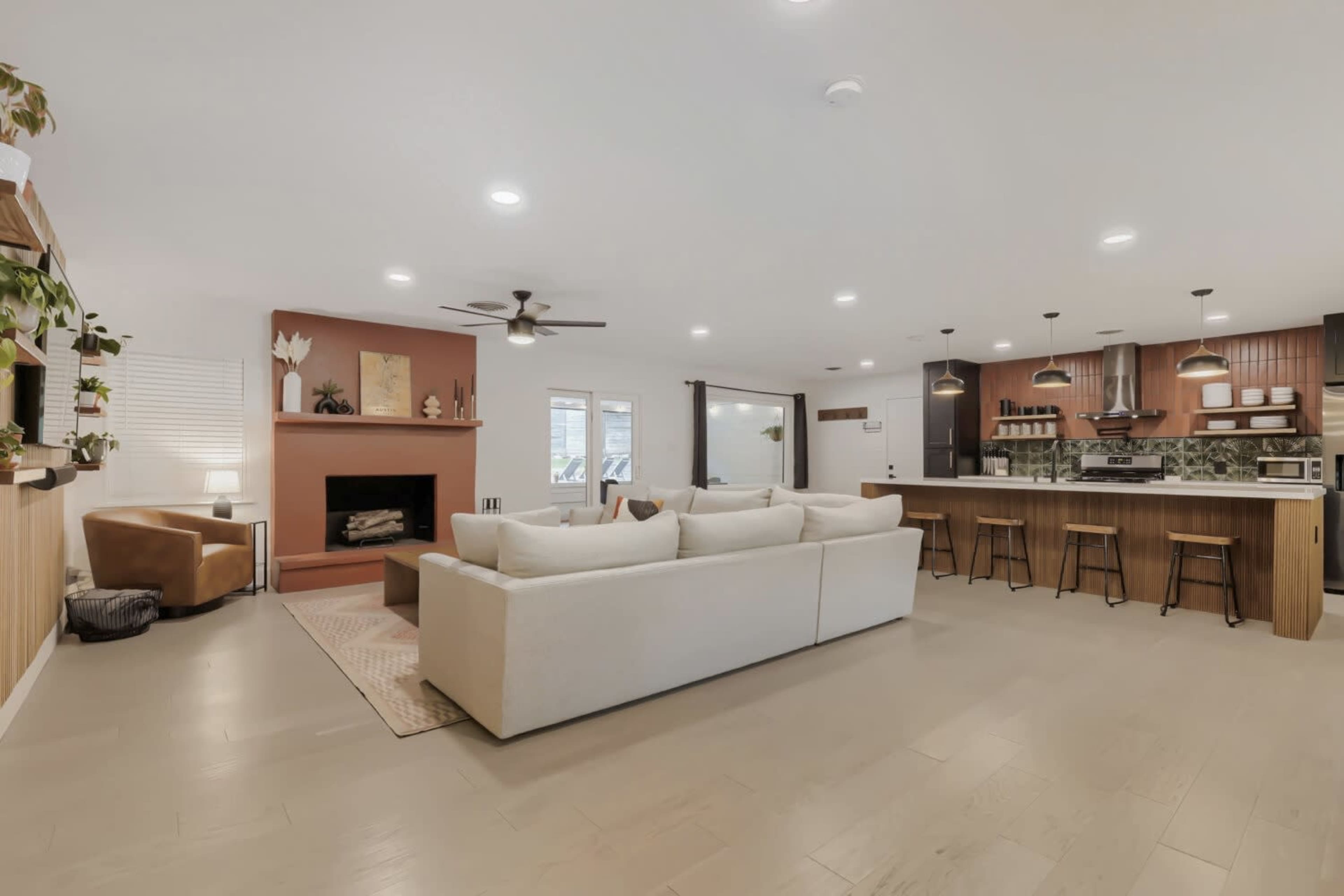 The image shows a modern living room with a sectional sofa facing a fireplace, alongside an open kitchen featuring bar stools and cabinetry.