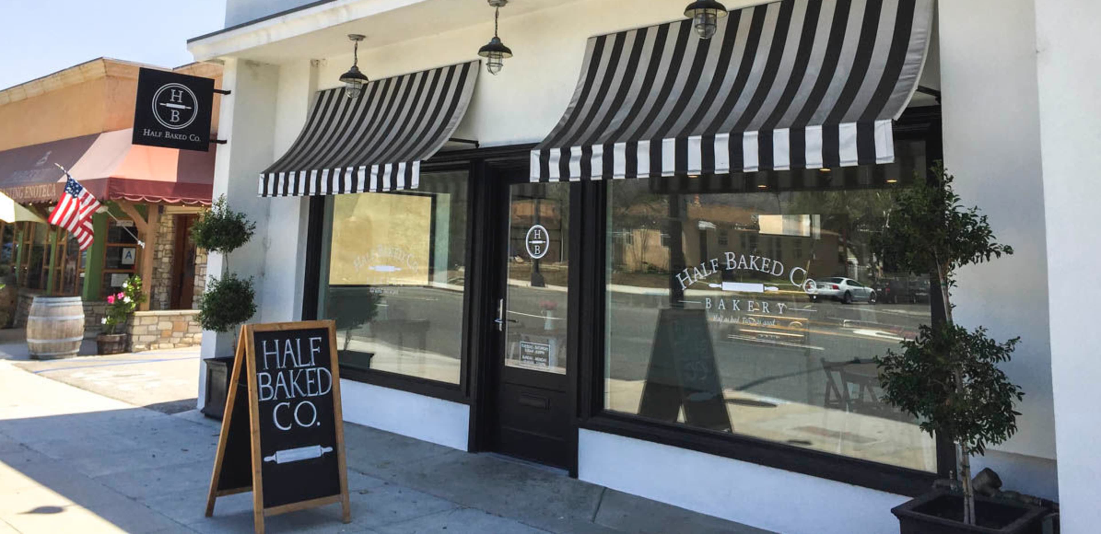 The exterior of a bakery named "Half Baked Co." features large windows with black and white striped awnings and a sidewalk sign.