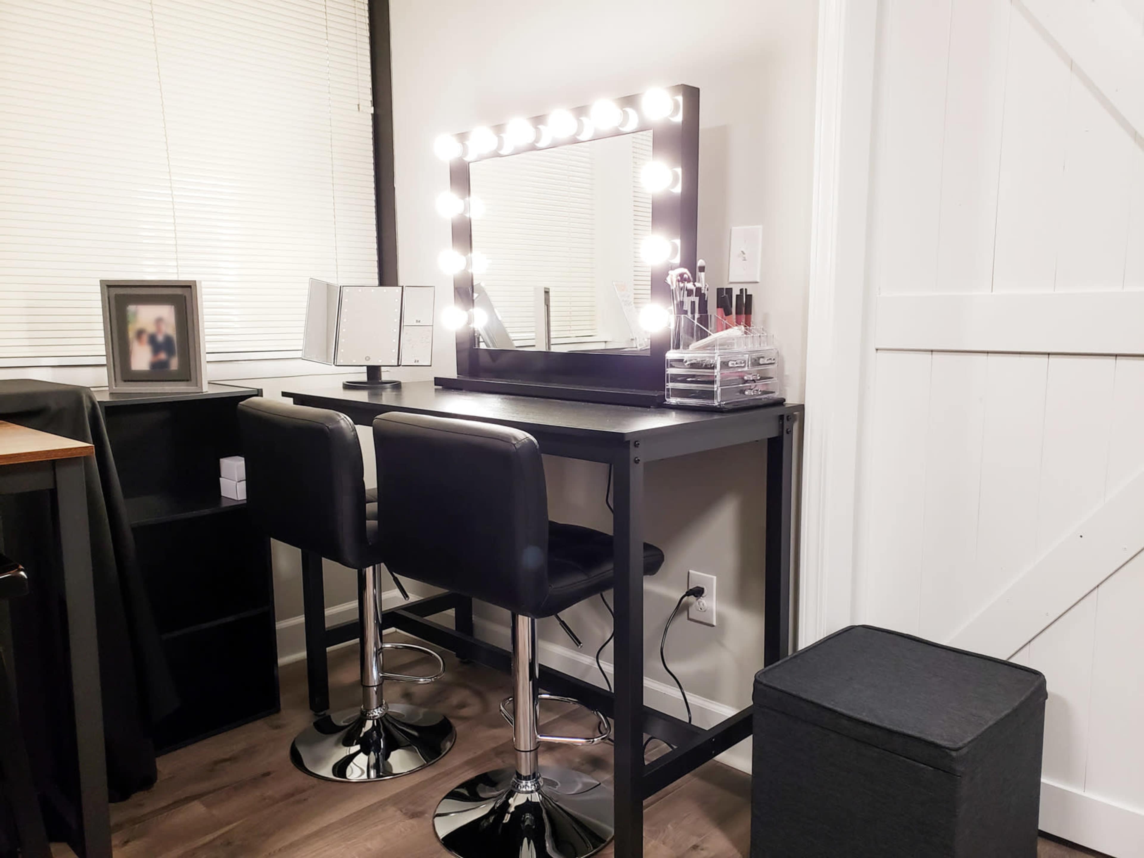 A modern vanity area features a mirror surrounded by lights, two black bar stools, and storage units in a well-lit room.