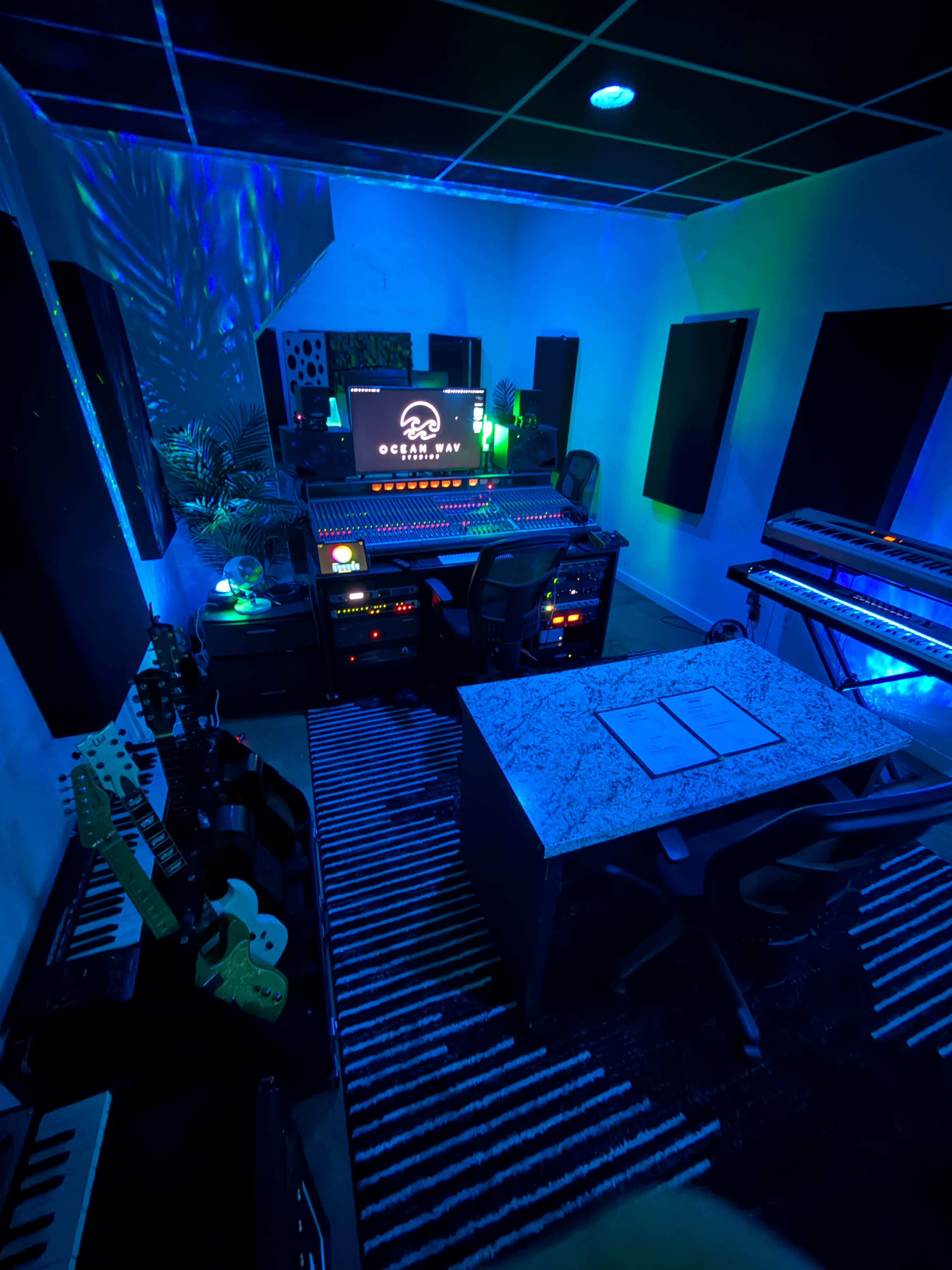 The image shows a music production studio with a large mixing console, computer setup, musical instruments, and ambient lighting.