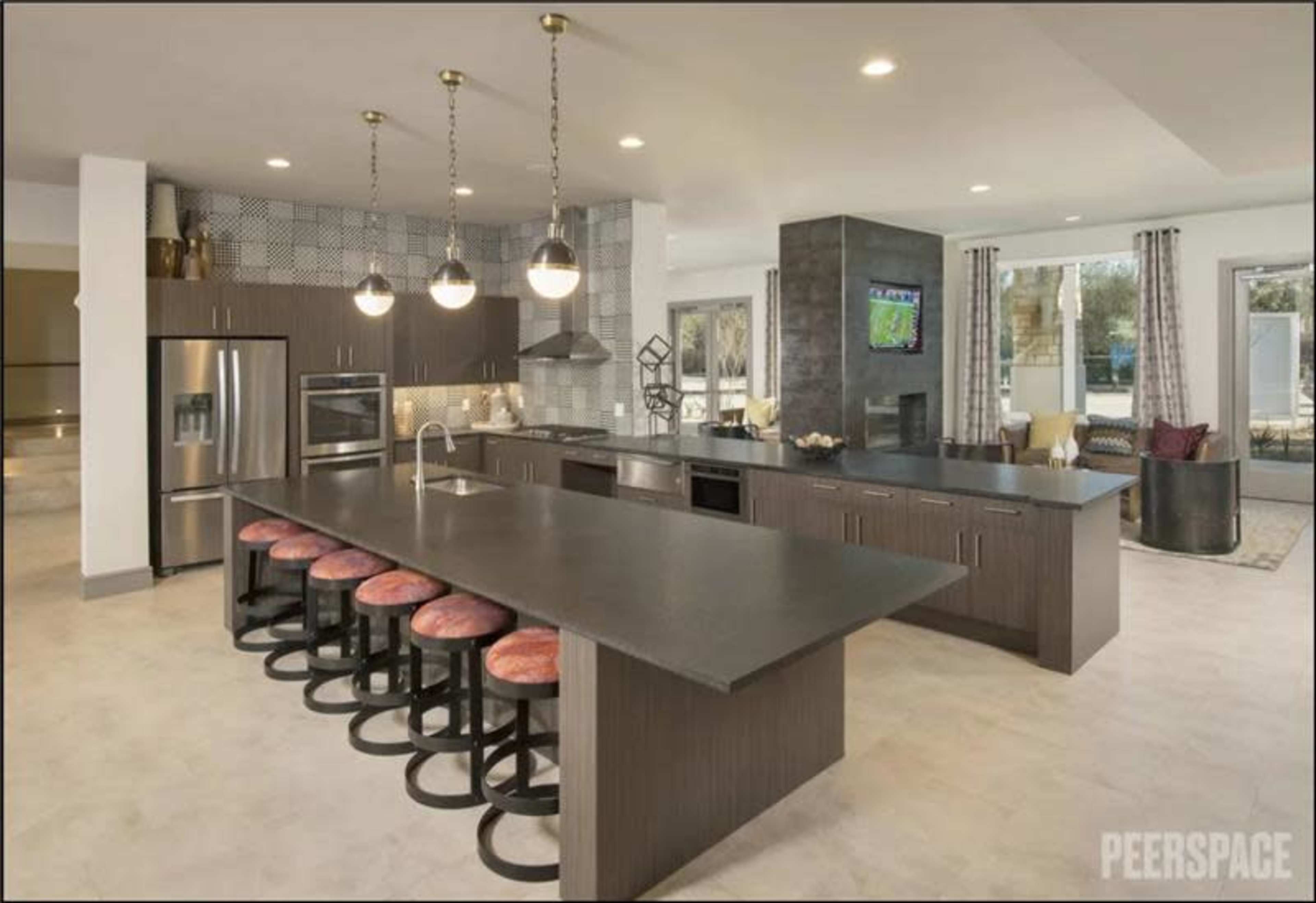 The image shows a modern kitchen with an expansive dark countertop island, bar stools, stainless steel appliances, and large windows that provide natural light.