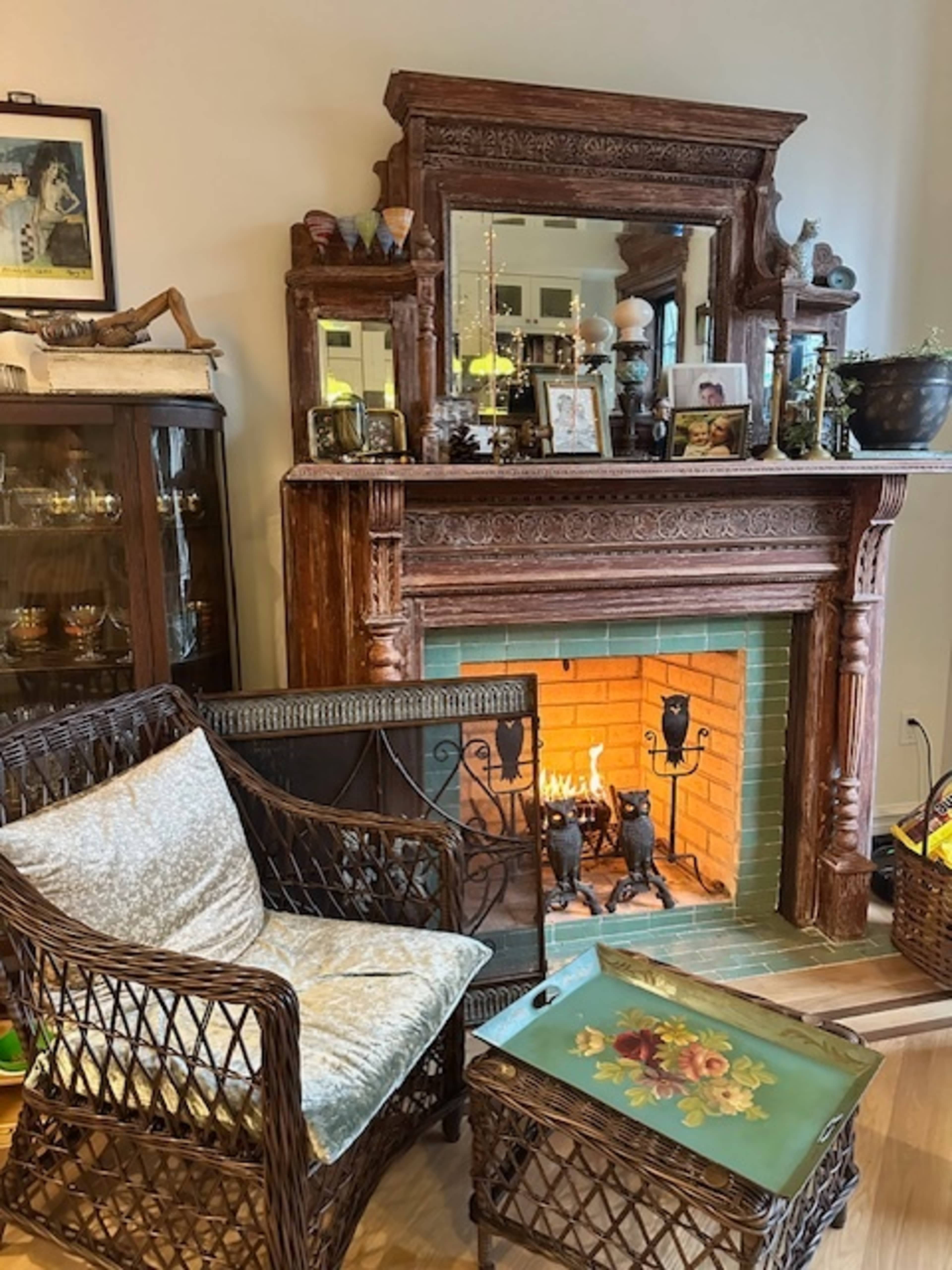 The image shows a cozy living room with a vintage wooden fireplace adorned with a mirror and decorations, accompanied by a wicker chair and small table.