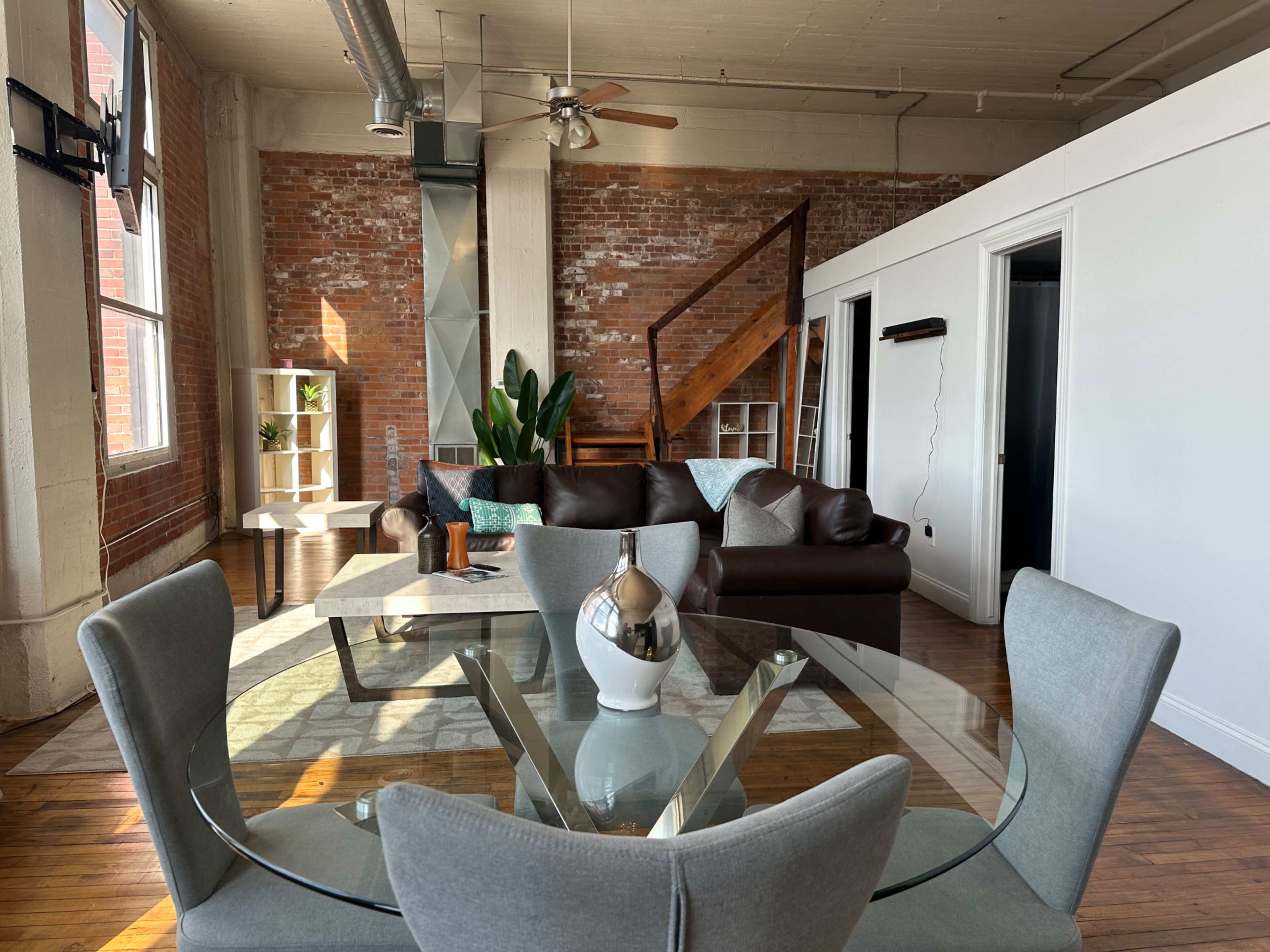 The image shows a modern, open-concept living space featuring exposed brick walls, a sofa, and a dining table set beneath natural light.