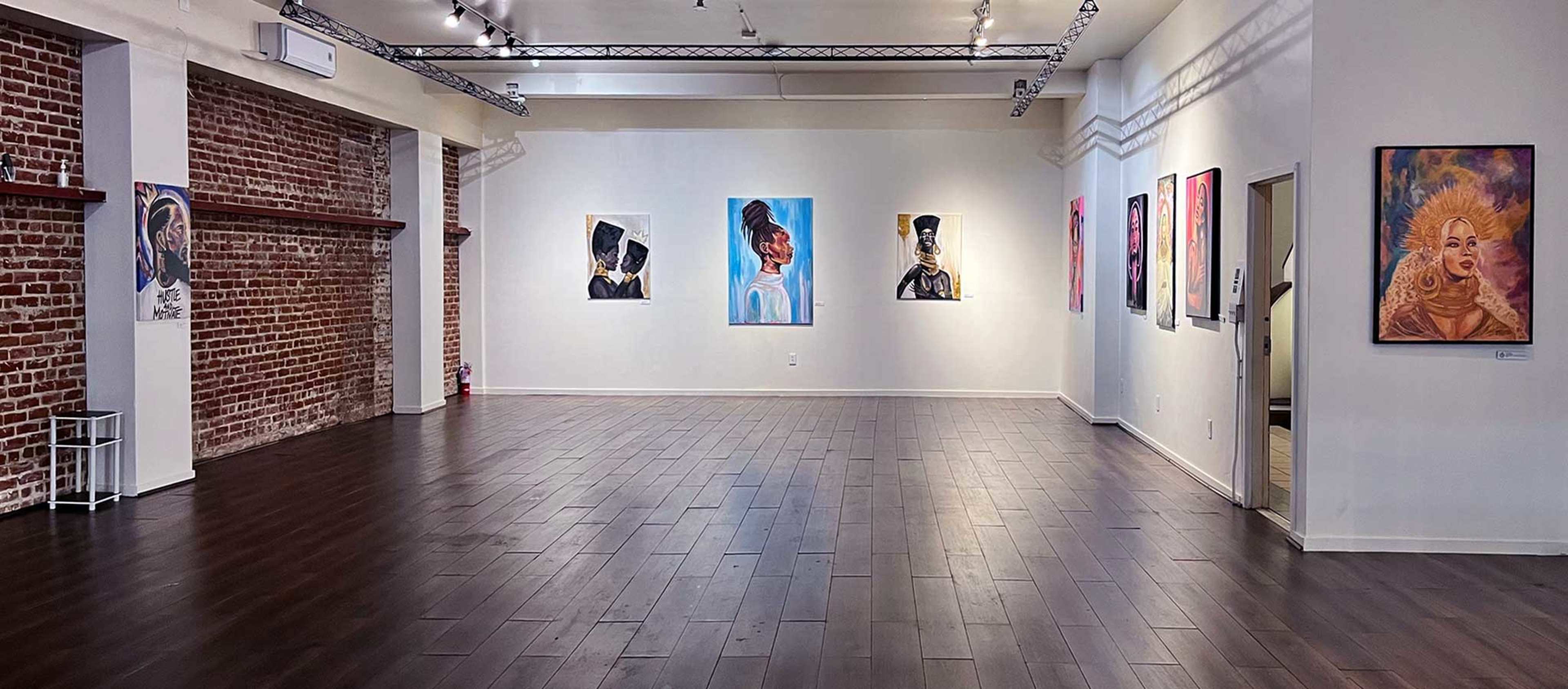 A spacious art gallery featuring various paintings displayed on the white walls, with exposed brick on one side and polished wooden flooring.