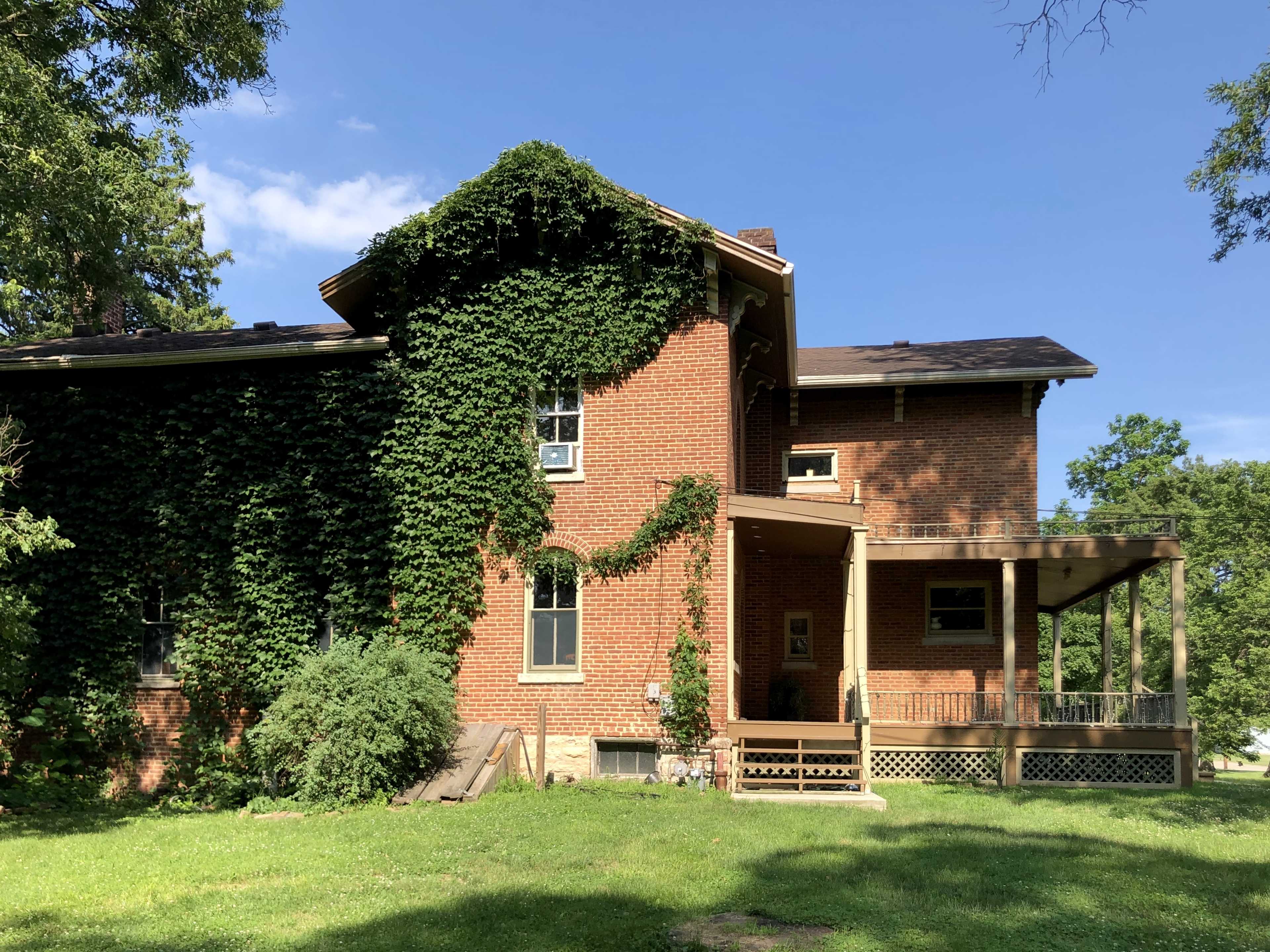 A two-story brick house enveloped in green ivy features a covered porch and is surrounded by tall grass and trees.