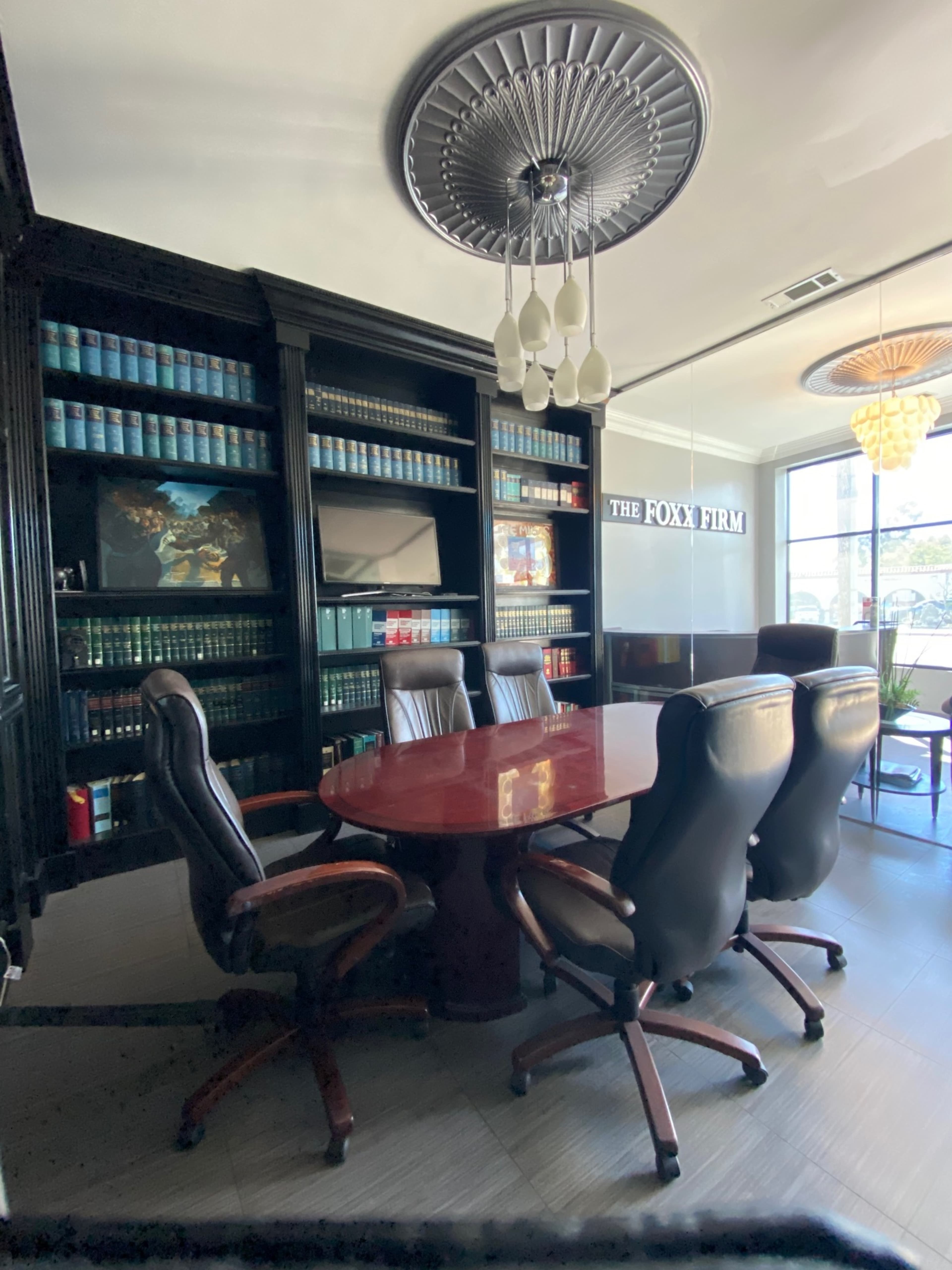 A conference room features a round table surrounded by black leather chairs, with a bookshelf filled with green law books in the background.