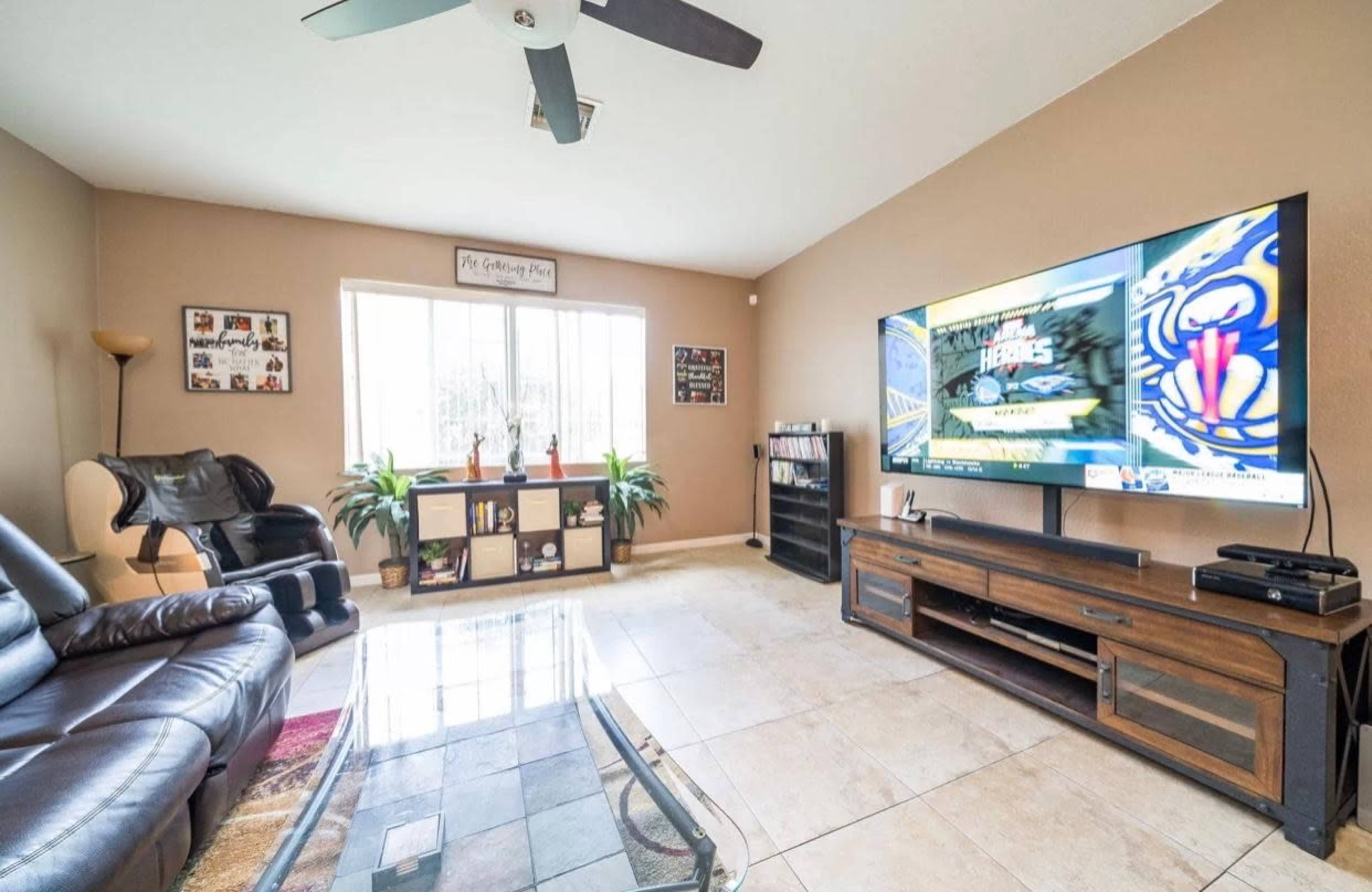 A living room features a leather sofa, a coffee table, a large television on an entertainment unit, and a window with natural light.