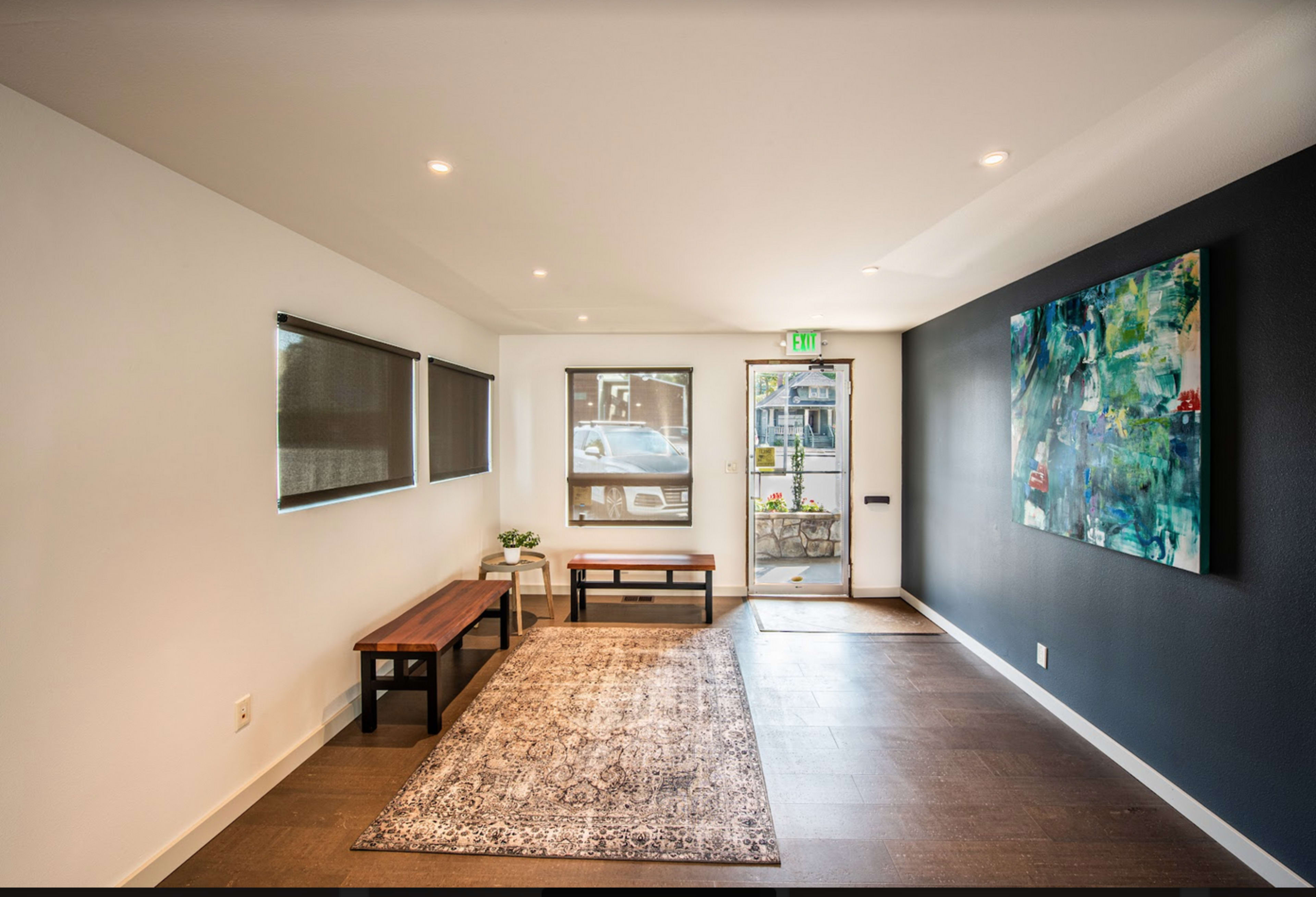 The image shows a modern entryway with a rug, two benches, and a large abstract painting on the wall, leading to a front door with a view of an outdoor area.