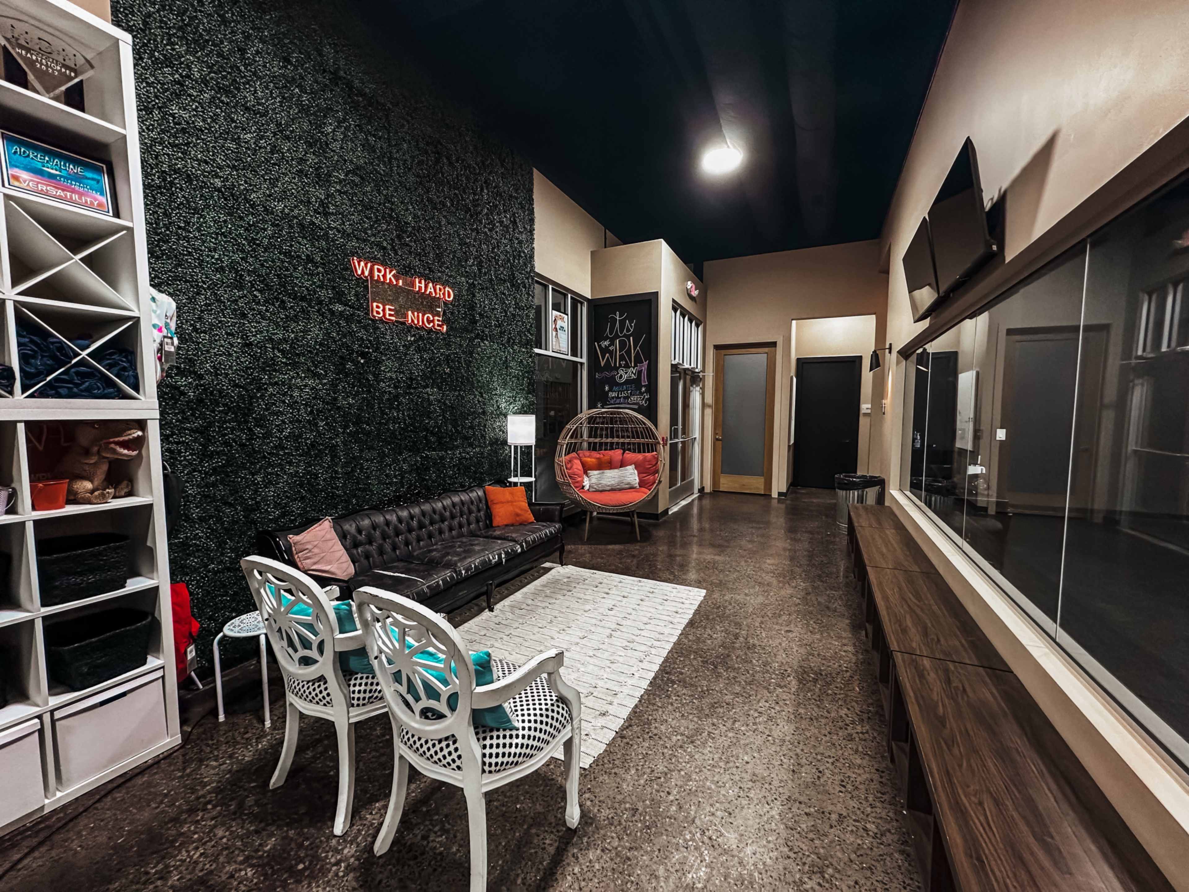 The image shows a cozy workspace with a green textured wall, a black leather couch, a hanging chair, and a large storage unit, all arranged in a modern, well-lit setting.