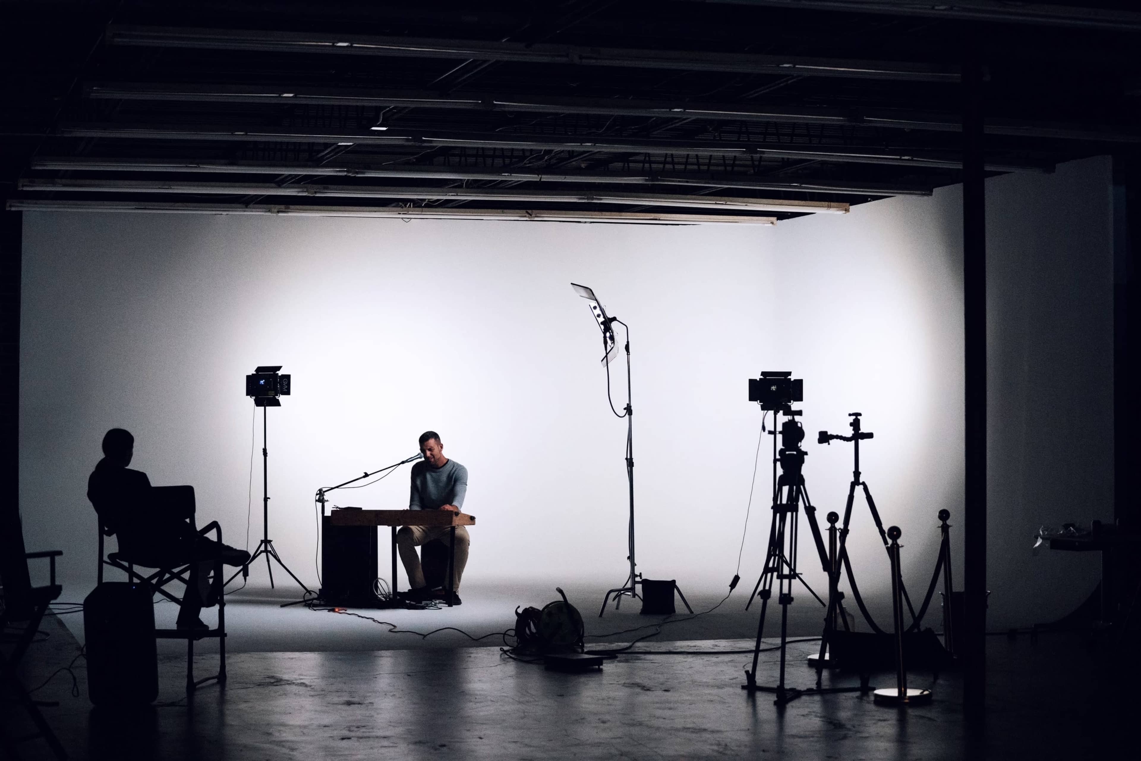 A musician sits at a table in a dimly lit studio with cameras and lighting equipment set up around him.