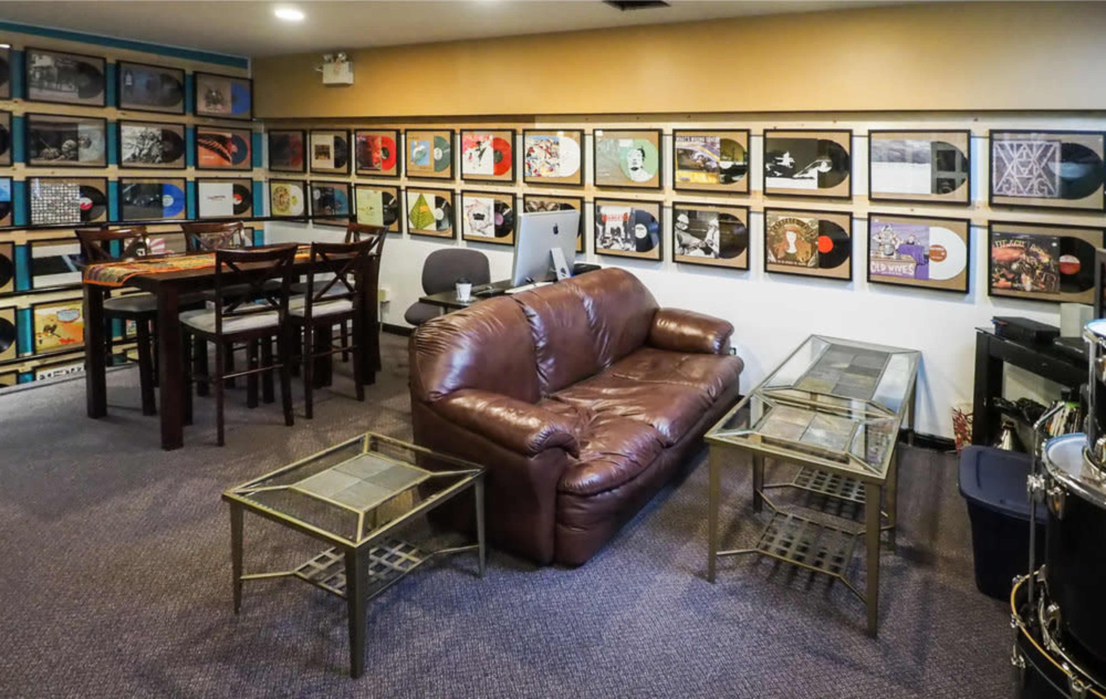 A room with a brown sofa and two glass-top tables, surrounded by framed album covers on the walls and a dining table in the corner.