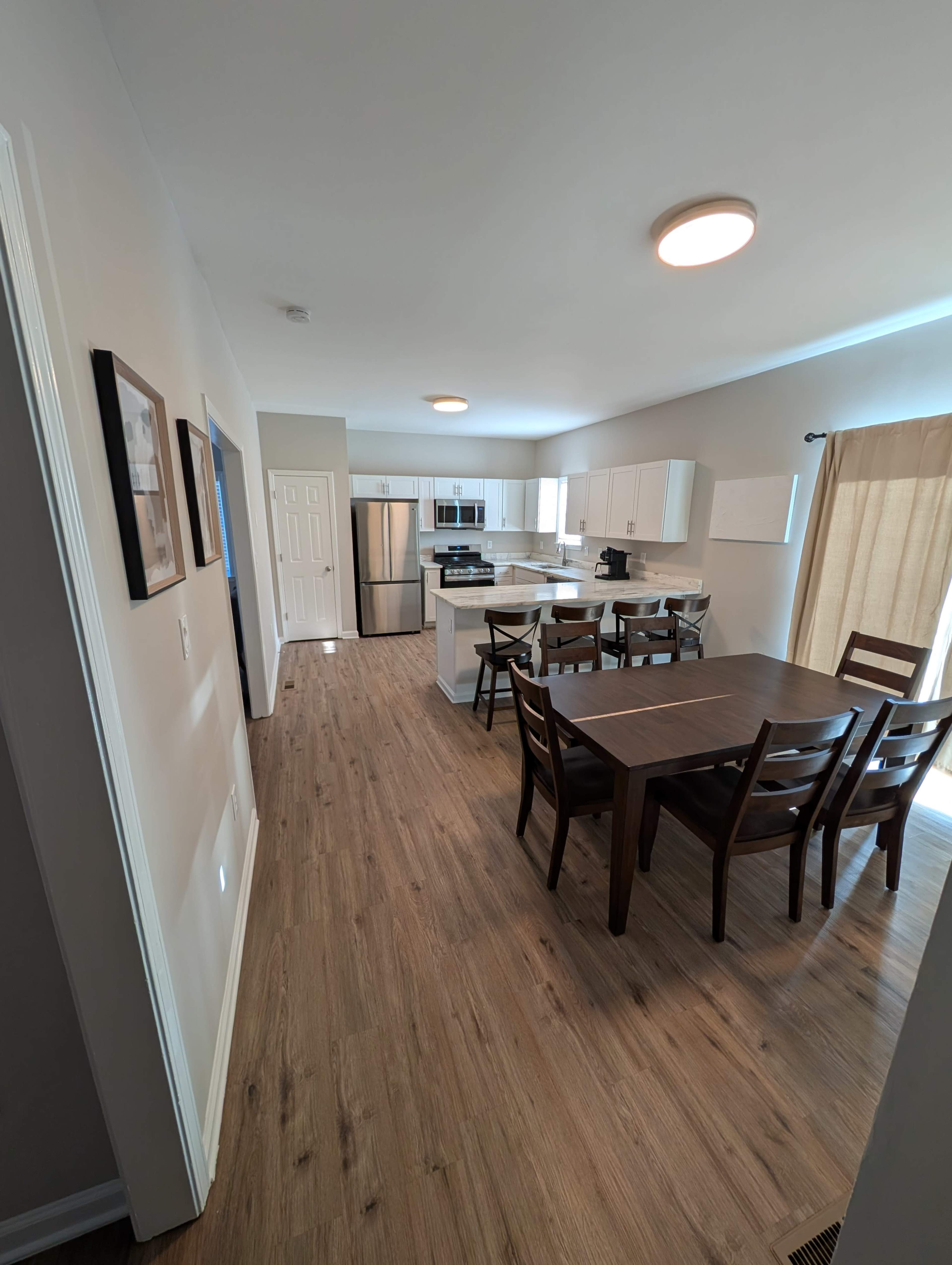 The image shows a modern kitchen and dining area with light wood flooring, white cabinets, and a dark wooden dining table surrounded by chairs.