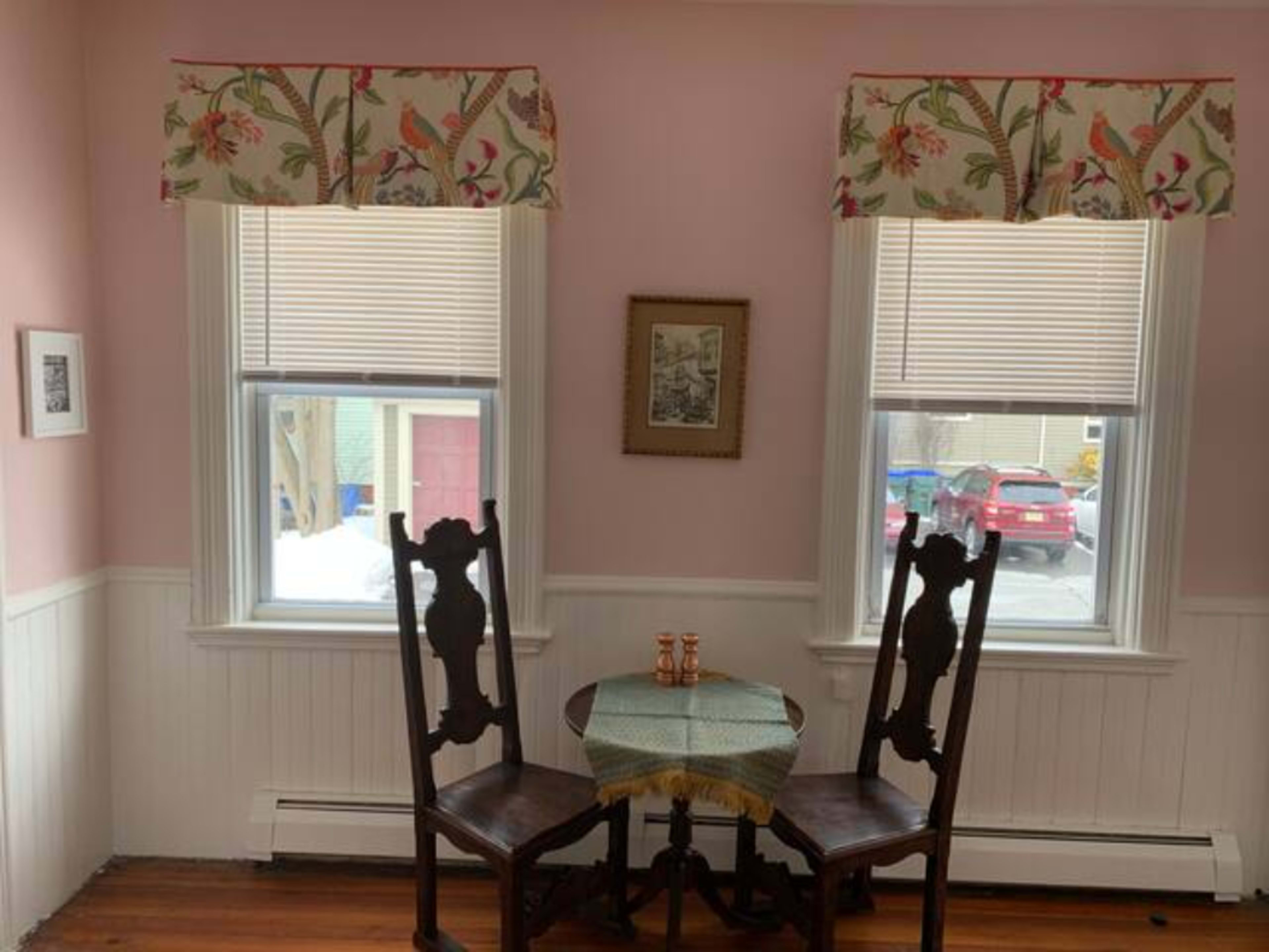 A small dining area with a round table and two wooden chairs, flanked by windows dressed with floral valances, and a framed picture on the wall.