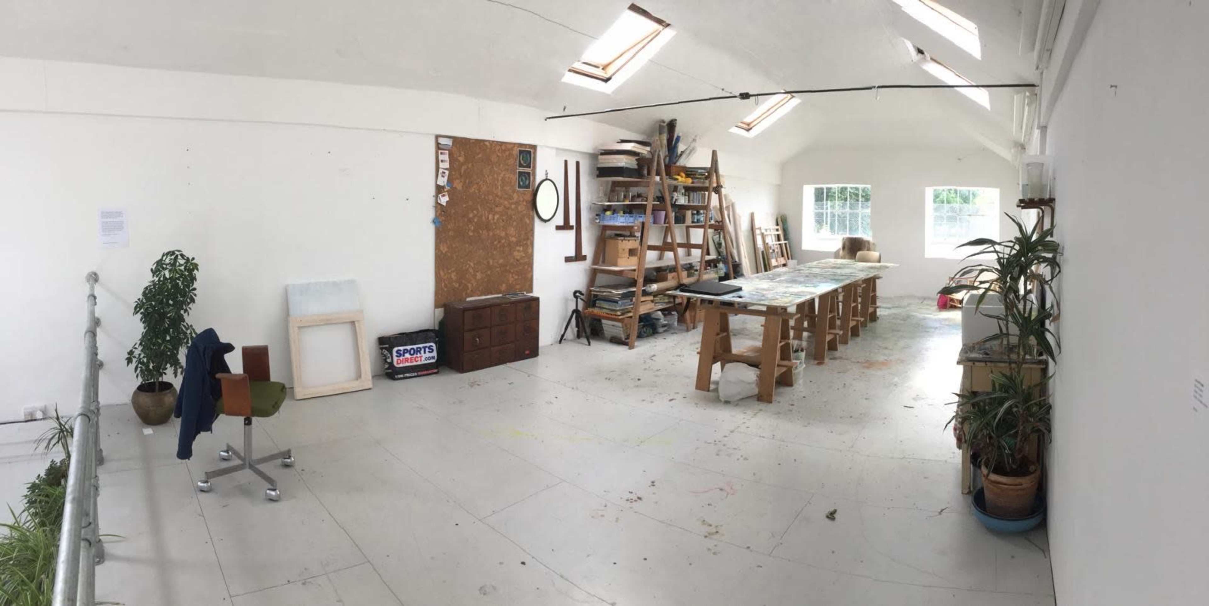 The image shows a spacious, well-lit art studio with a long wooden table, shelves filled with materials, and a few potted plants.
