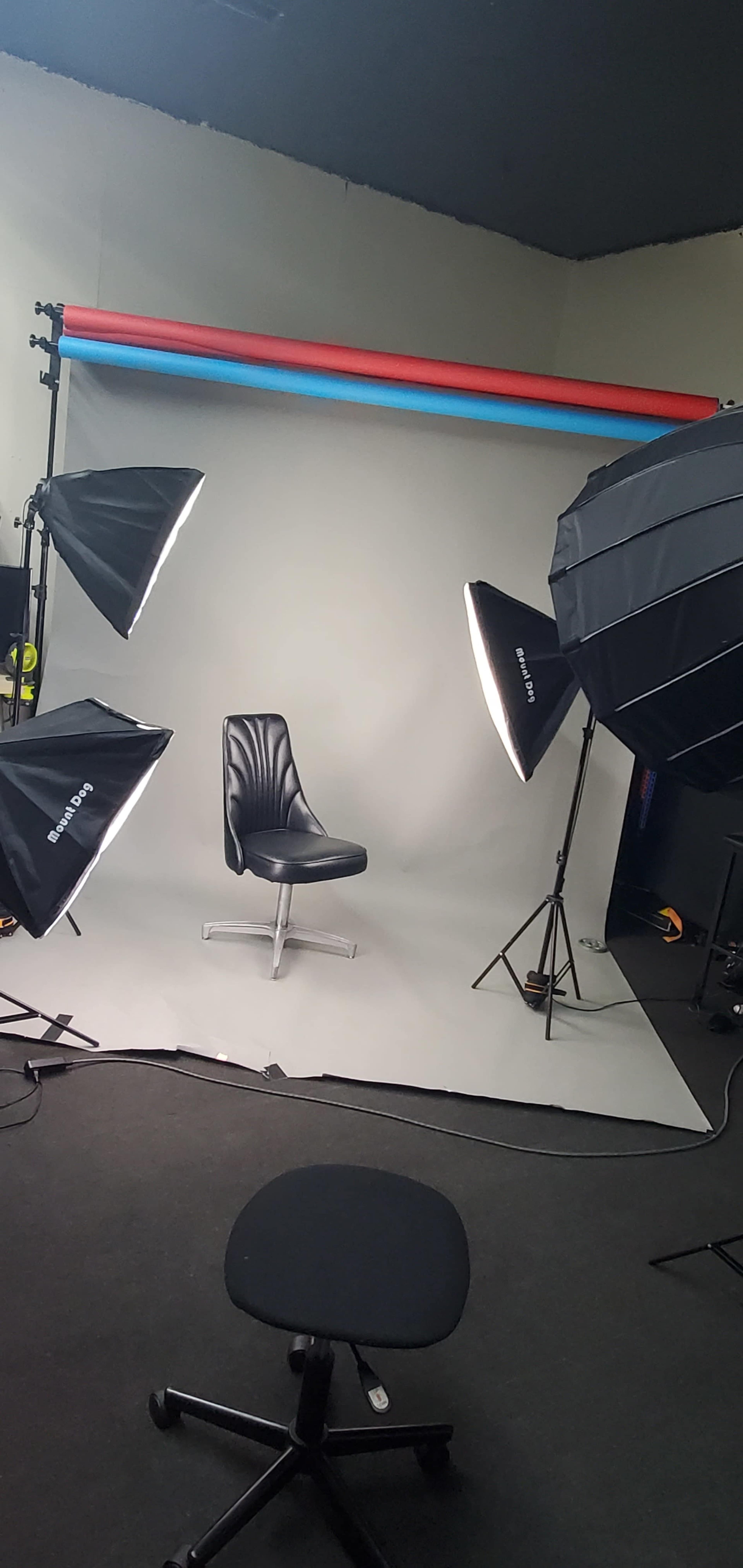 A photography studio setup with a black chair positioned on a gray floor, illuminated by several softbox lights against a backdrop of colored fabric.