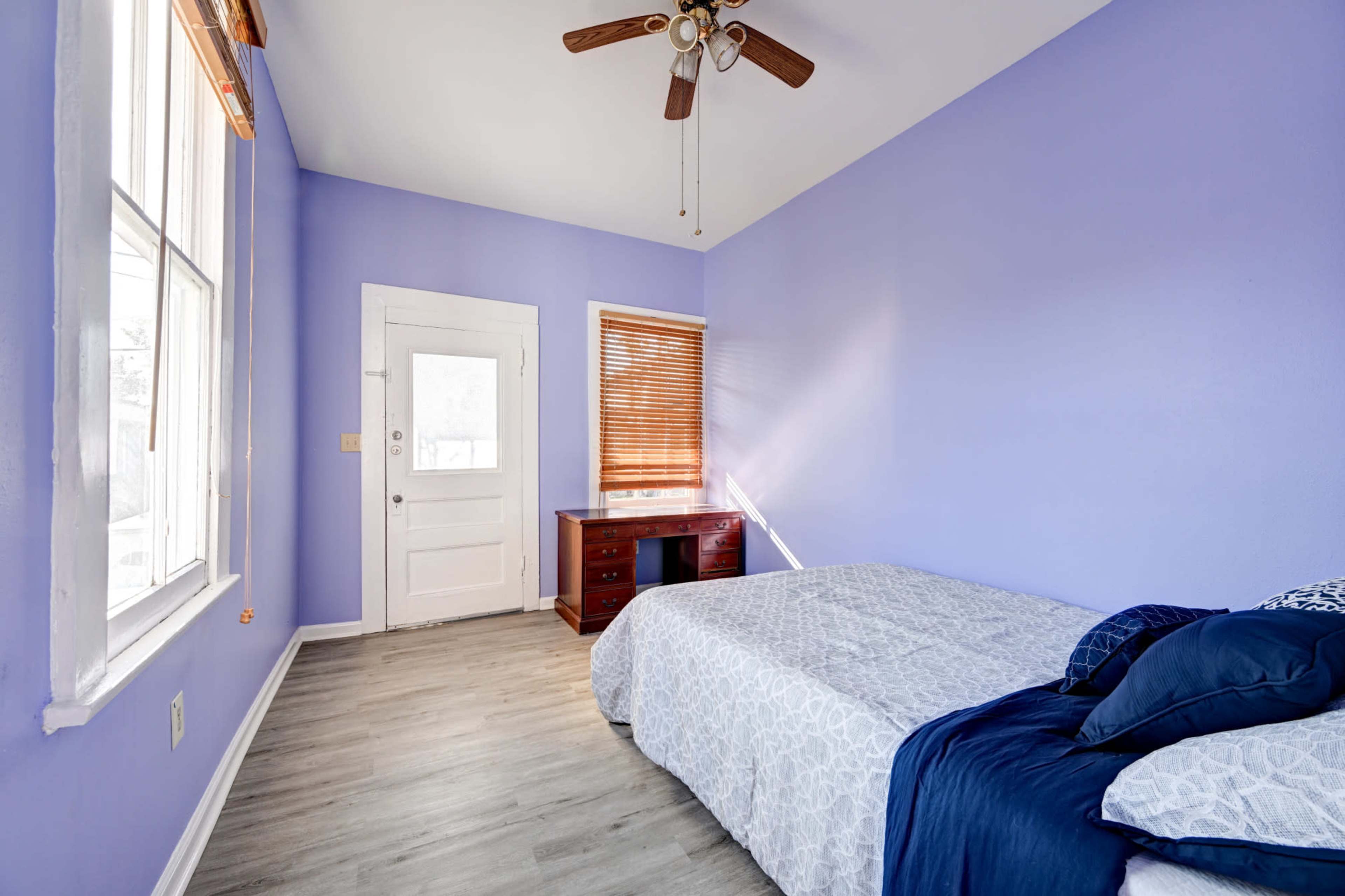 The room features a bed with a light-colored quilt against a purple wall, a wooden desk by a window with blinds, and a door leading outside.