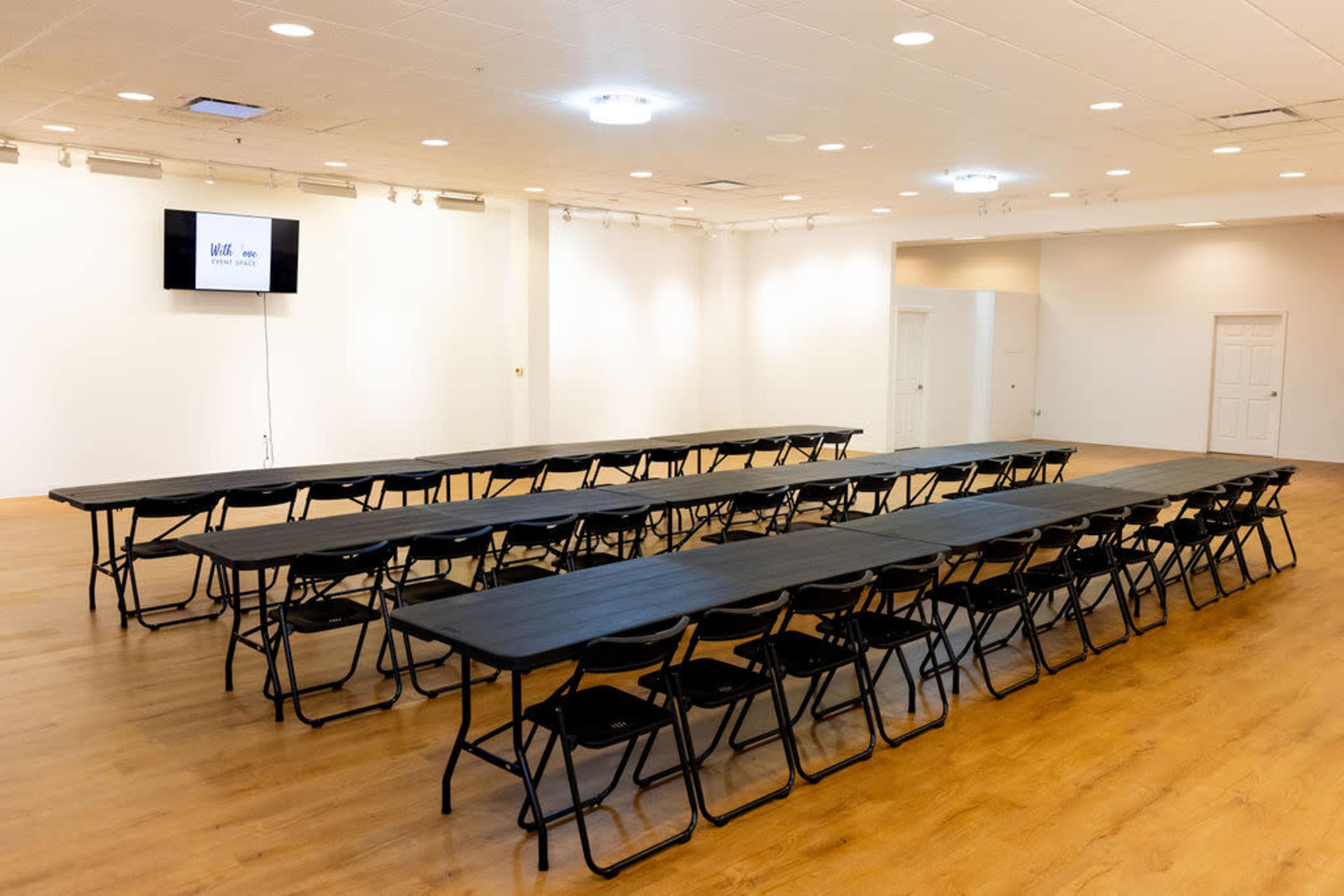 The image shows a large room set up with several black folding tables arranged in rows, accompanied by matching folding chairs, and features a mounted screen at one end.