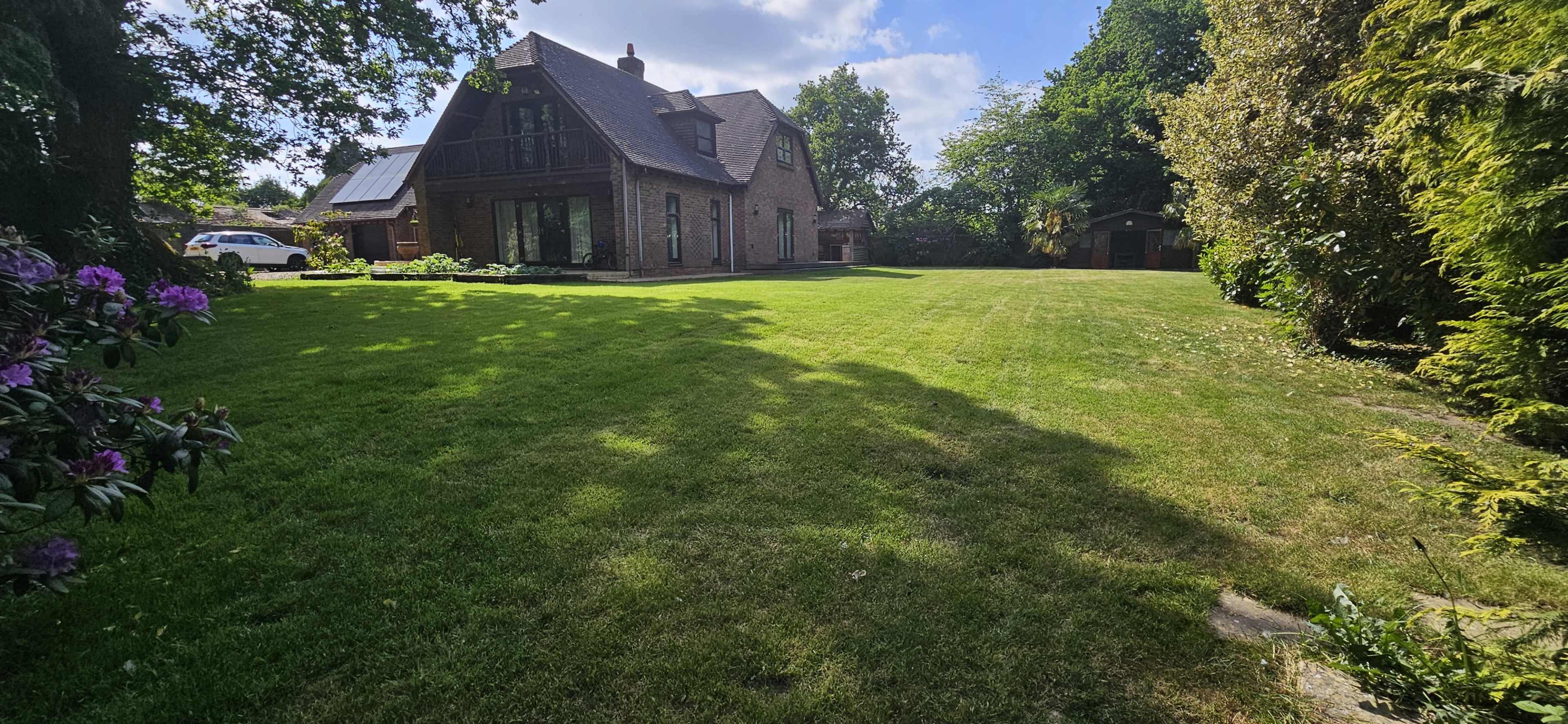The image shows a large, grassy yard in front of a modern, two-story brick house surrounded by trees and shrubs.
