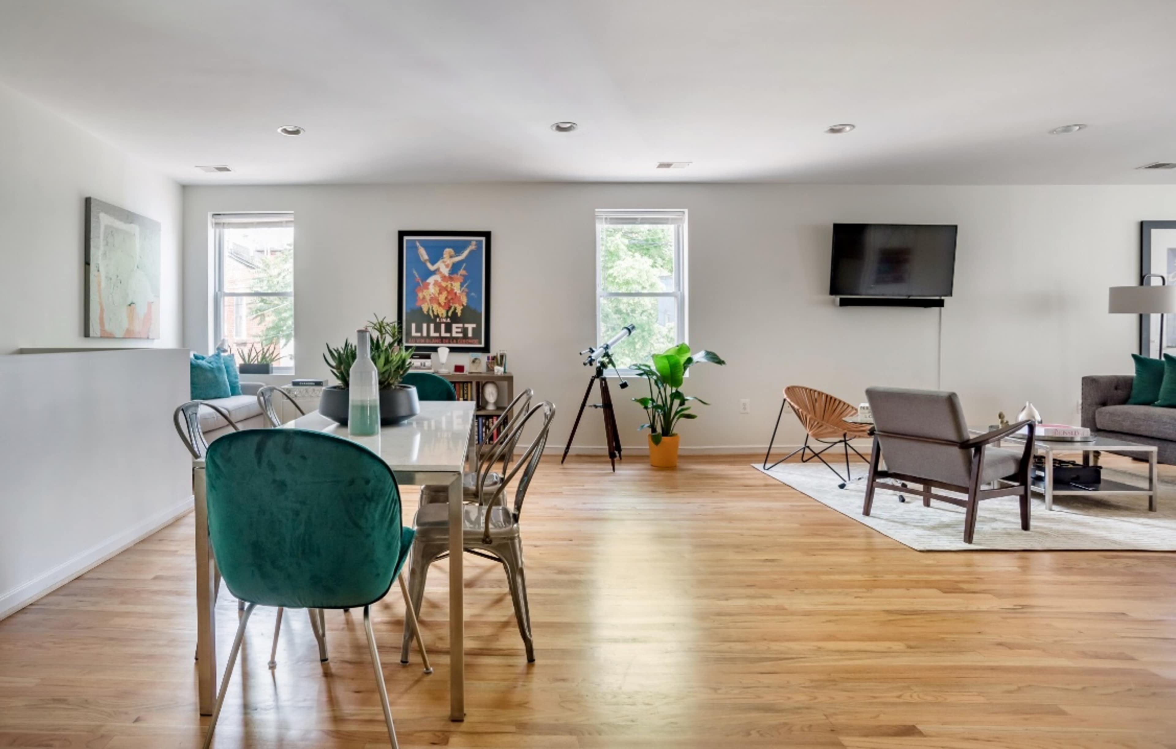 The image shows a modern living room and dining area with a wooden floor, a dining table, a couch, a TV, and decorative plants.