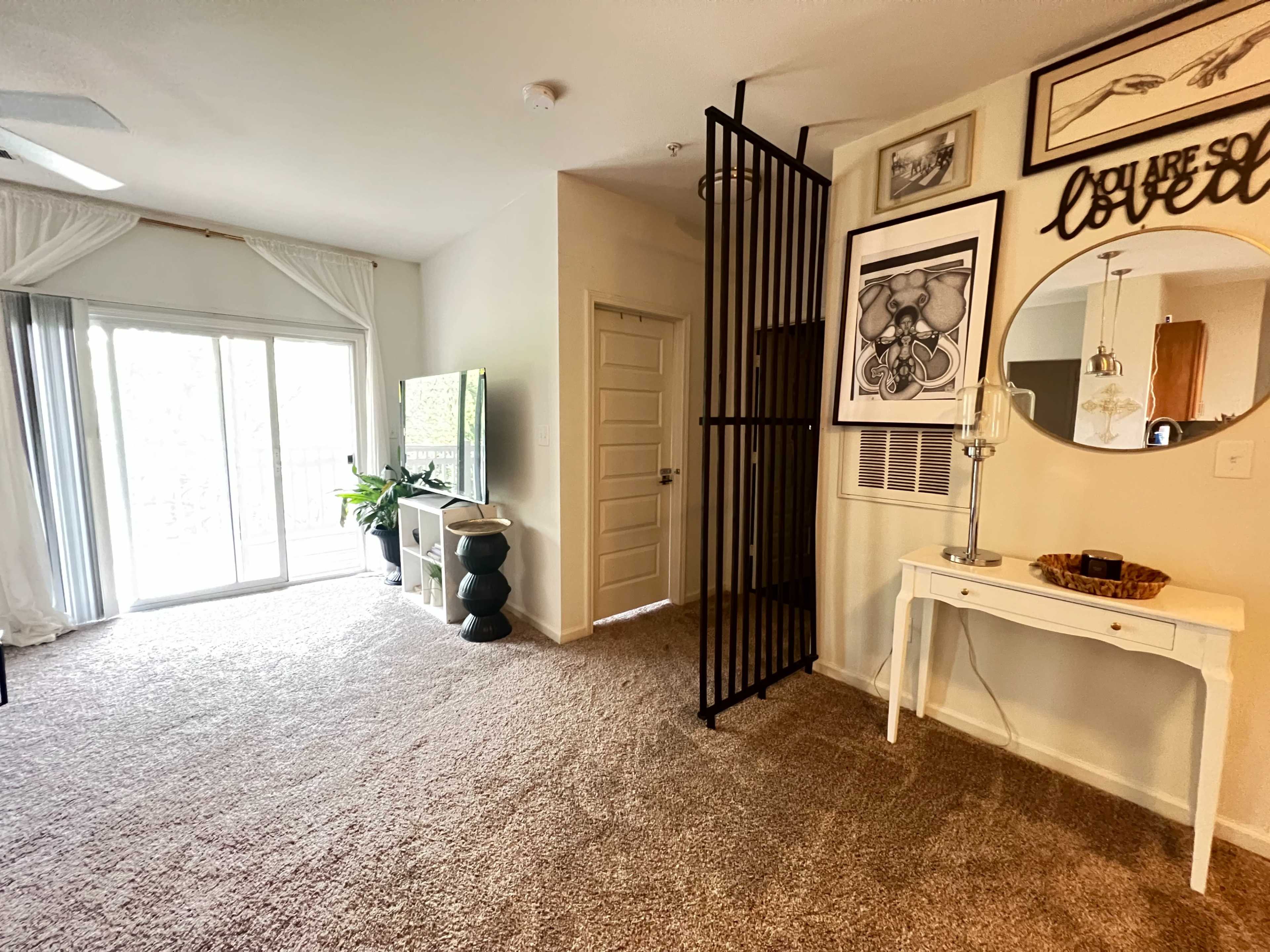 A living space with a carpeted floor, a television against the wall, a round mirror and table near the entrance, and sliding glass doors leading to a balcony.