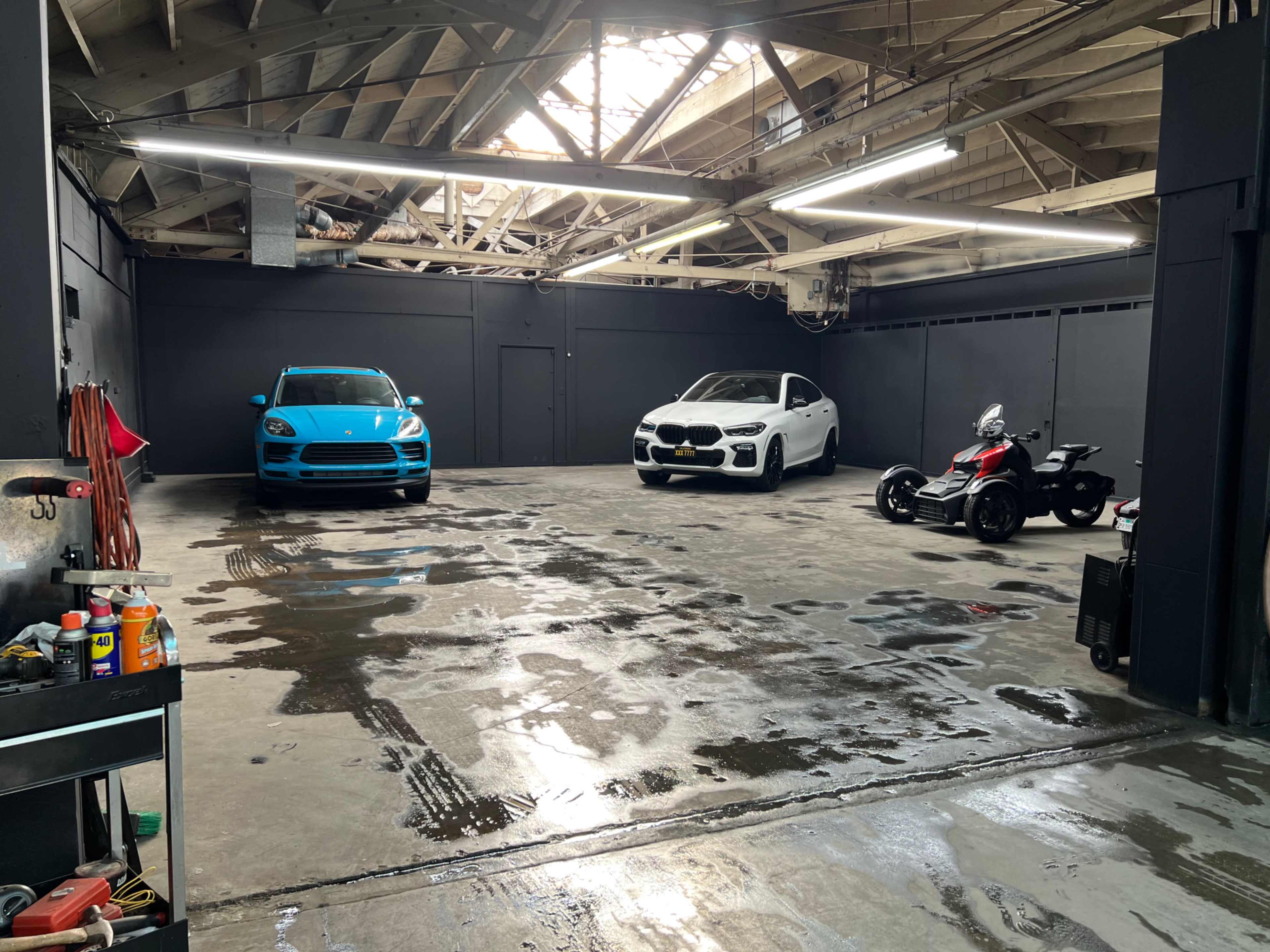 A spacious garage featuring a blue car, a white SUV, and a three-wheeled motorcycle, with a polished concrete floor and exposed beams overhead.