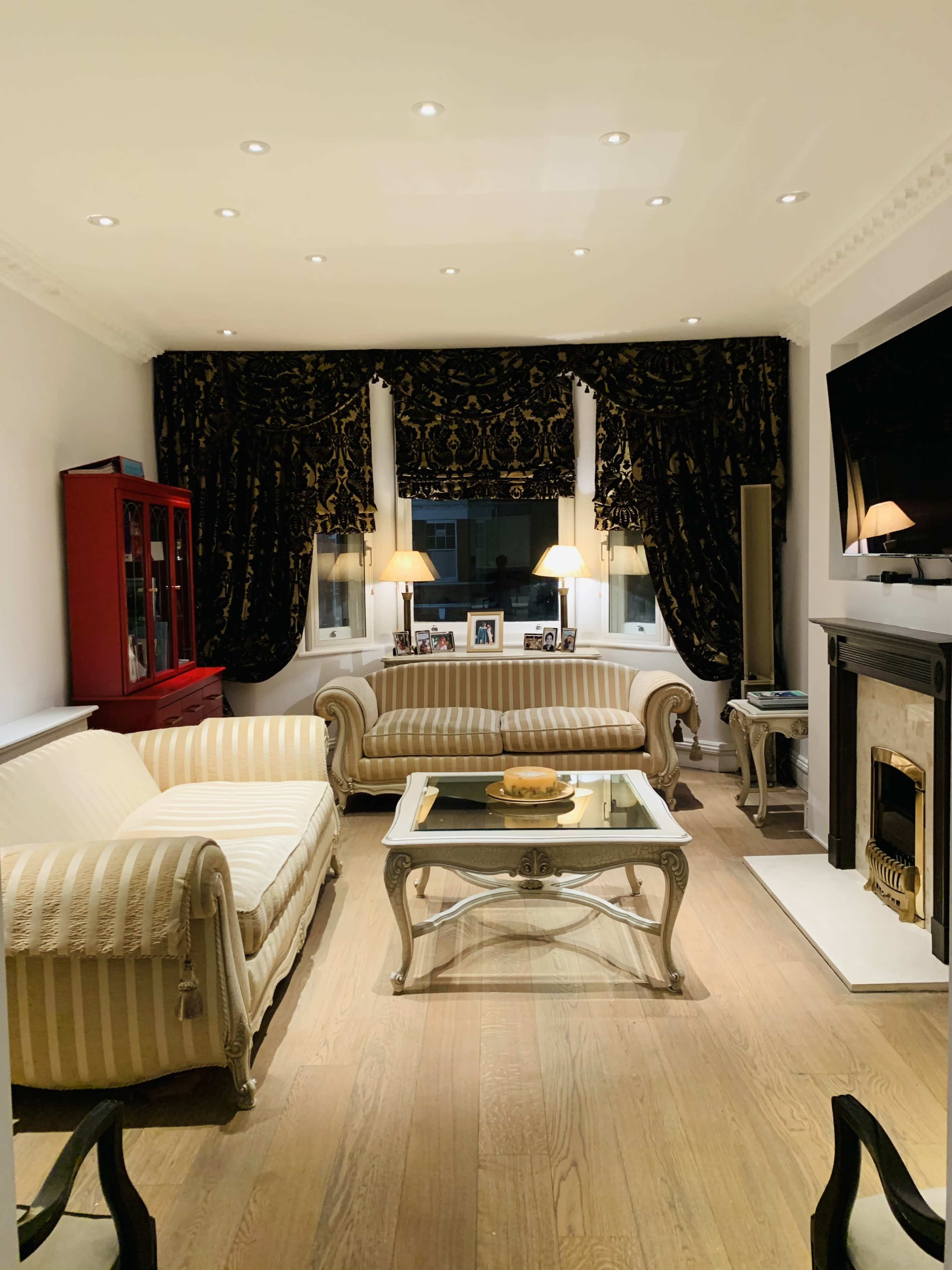 The living room features two striped sofas, a coffee table with a decorative centerpiece, and a fireplace, framed by dark curtains and illuminated by lamps.