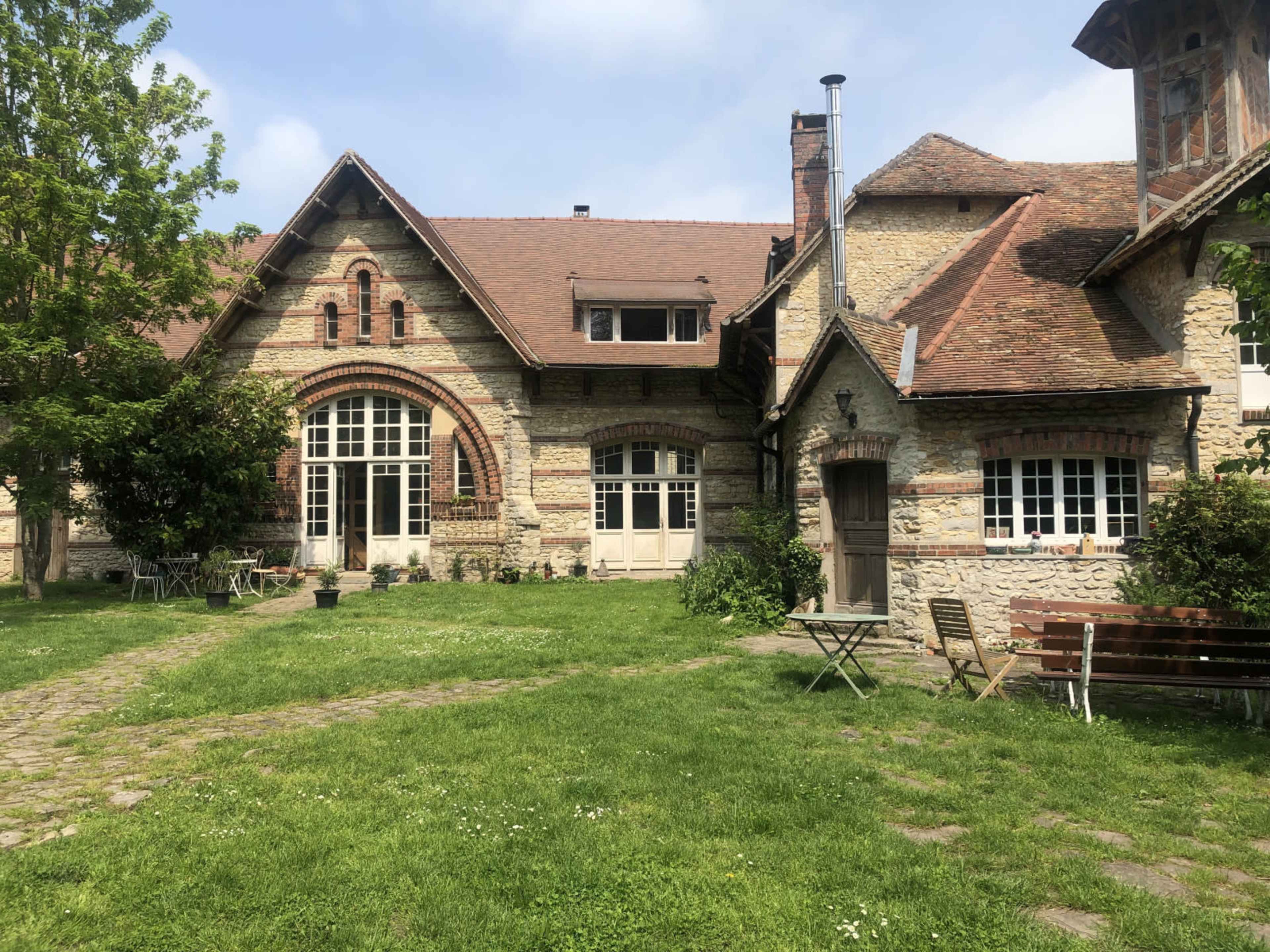 The image shows a rustic stone building with a sloped roof, surrounded by green grass and trees, featuring a pathway leading to a door.