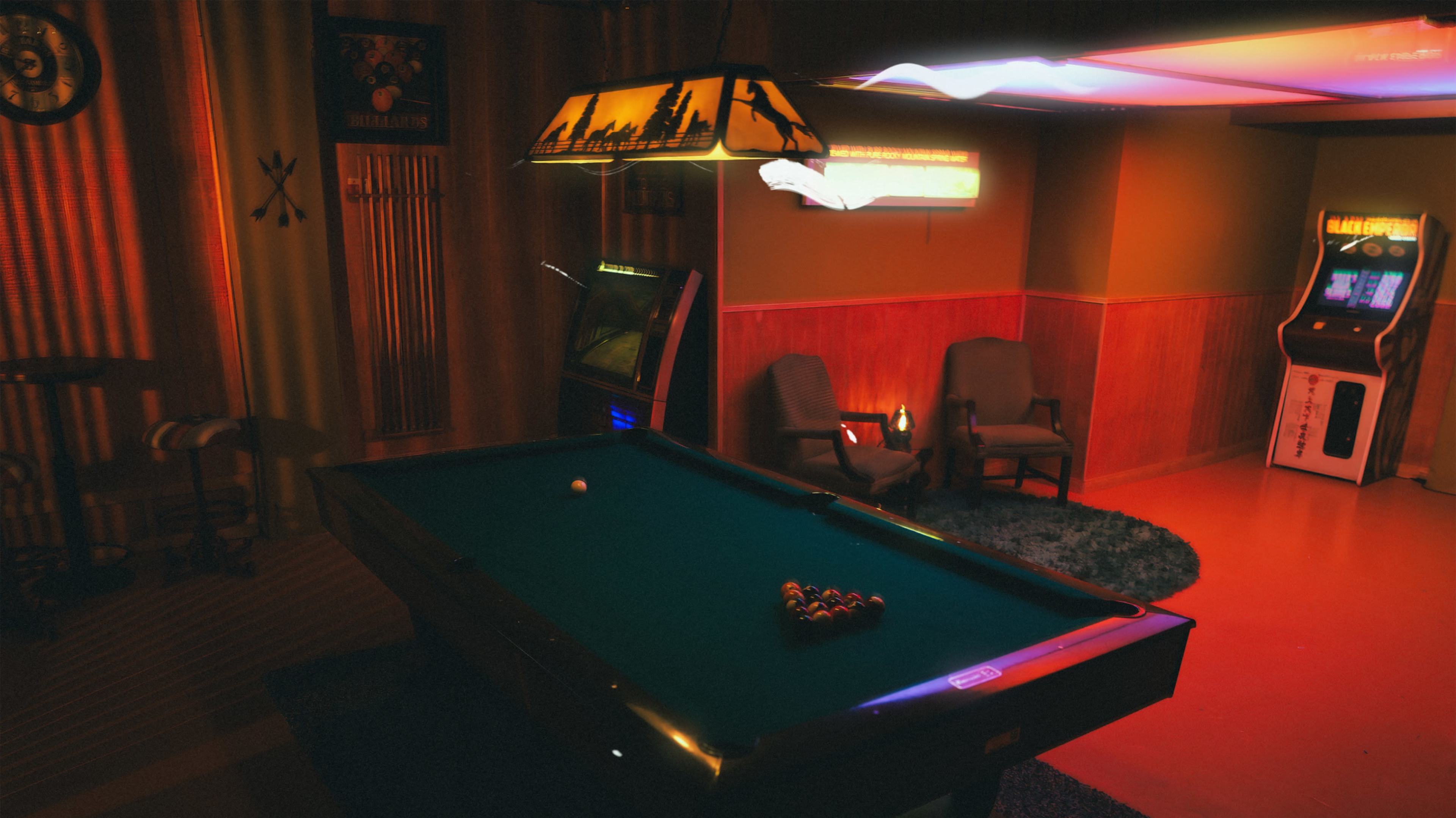 The image shows a dimly lit game room featuring a pool table with a triangle of balls, two chairs in the corner, and arcade machines against the wall.