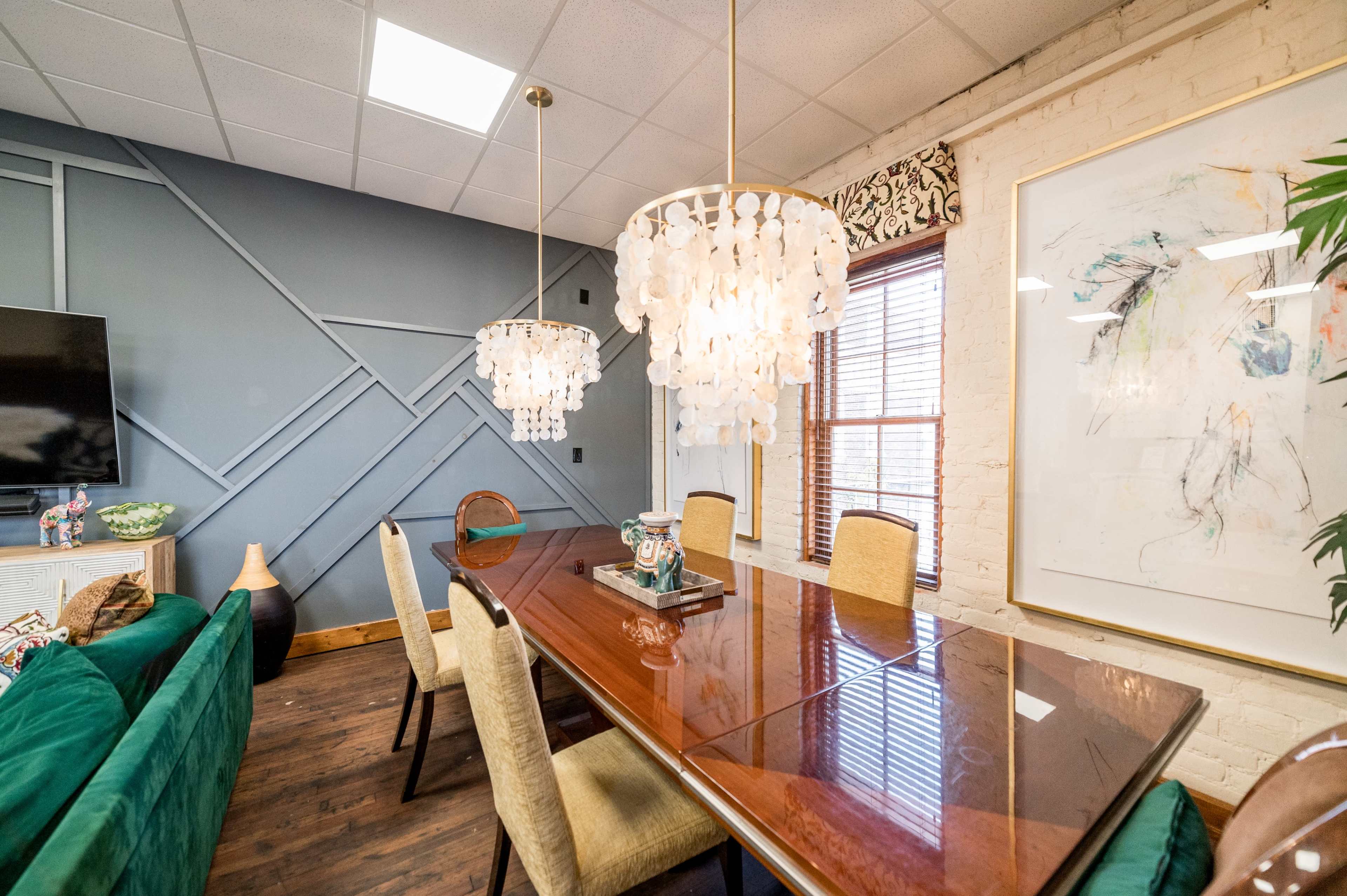 A modern conference room with a wooden table surrounded by upholstered chairs, two decorative light fixtures hanging above, and a large wall art piece alongside a television.