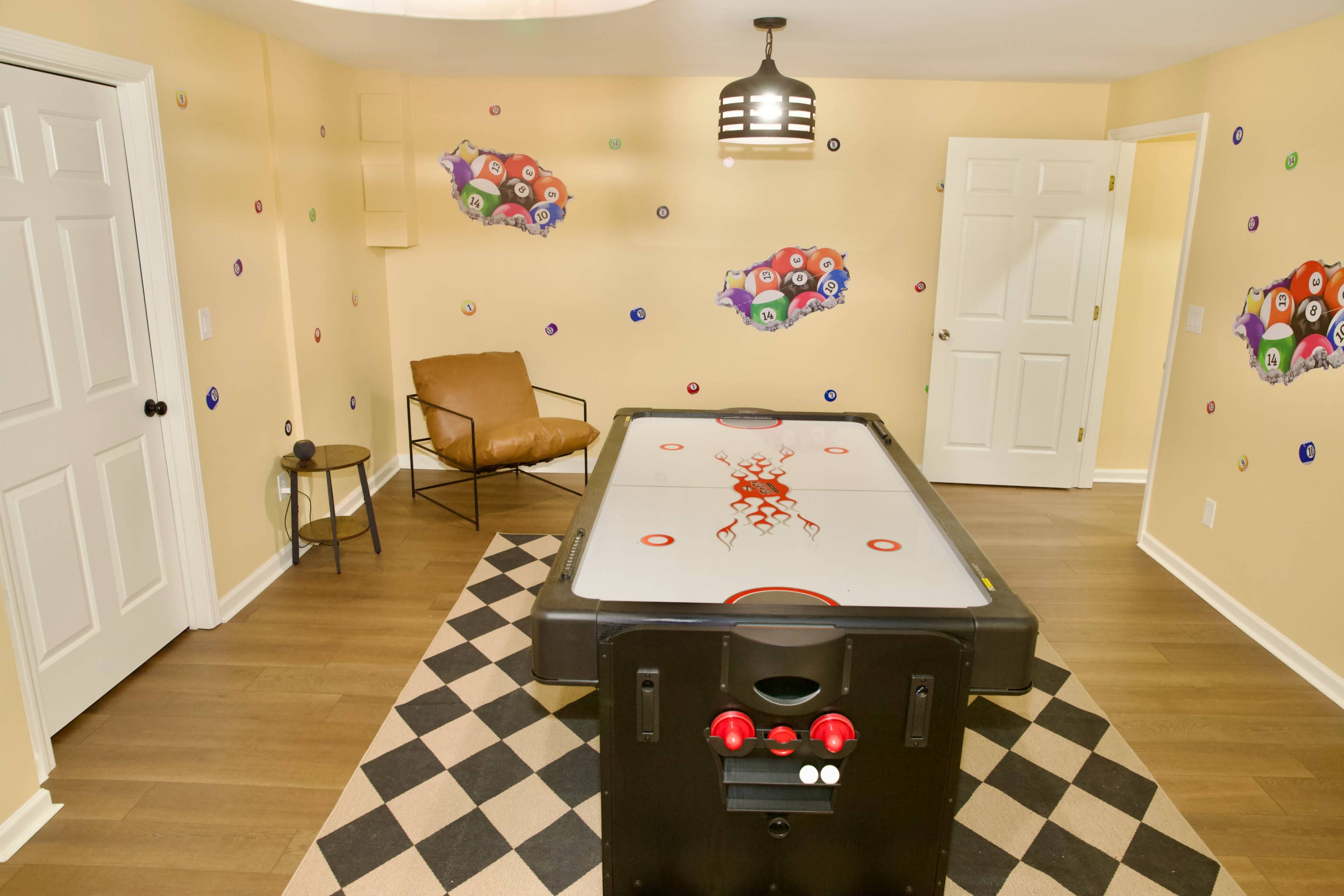 The image shows a game room featuring an air hockey table surrounded by light-colored walls adorned with decorative stickers and a brown armchair in the corner.