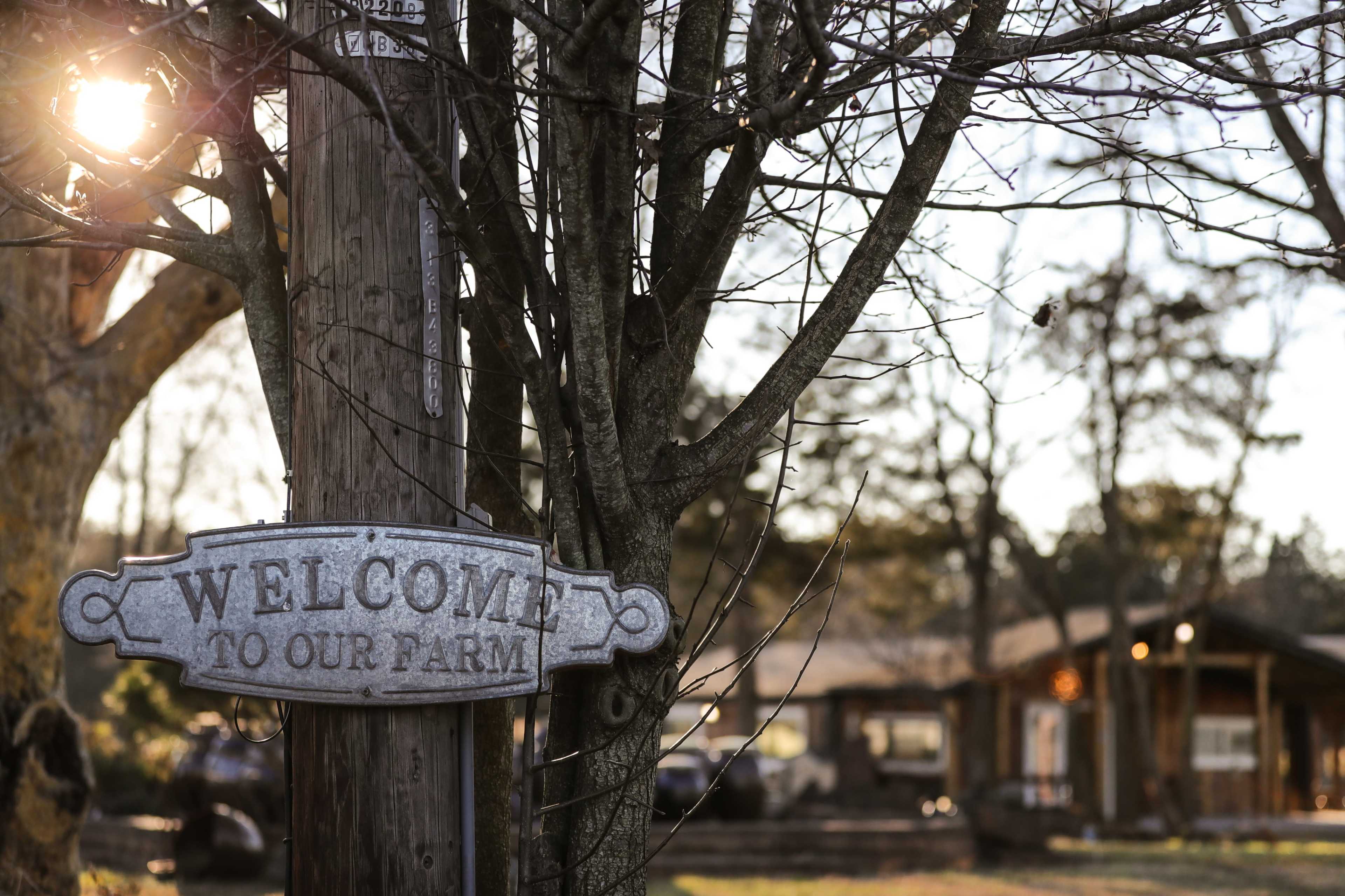 A metal sign reading "Welcome to Our Farm" hangs from a post in front of a rustic farmhouse surrounded by bare trees and sunlight filtering through the branches.