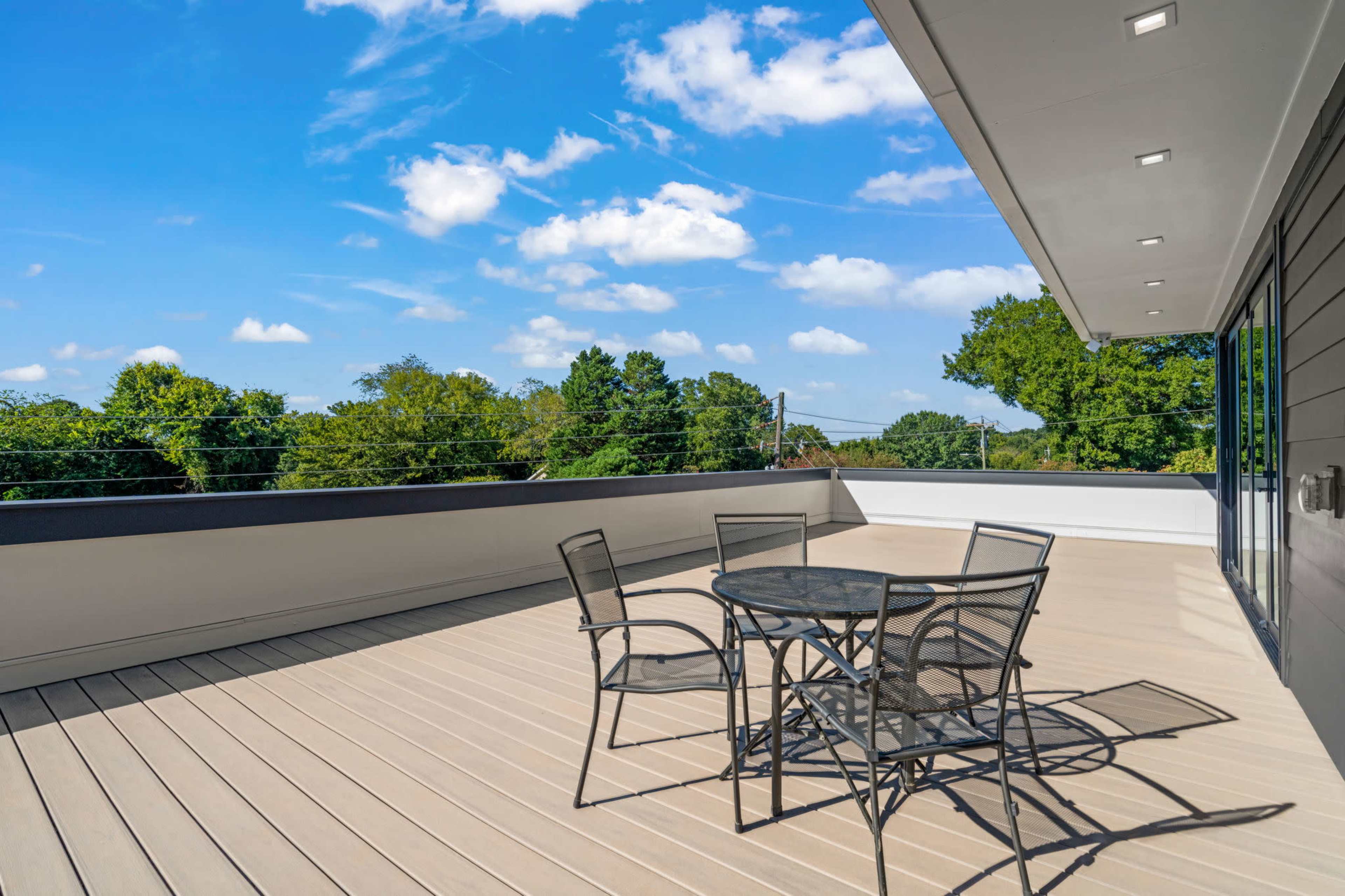 A spacious balcony features a round table with four chairs and overlooks green trees under a blue sky with scattered clouds.