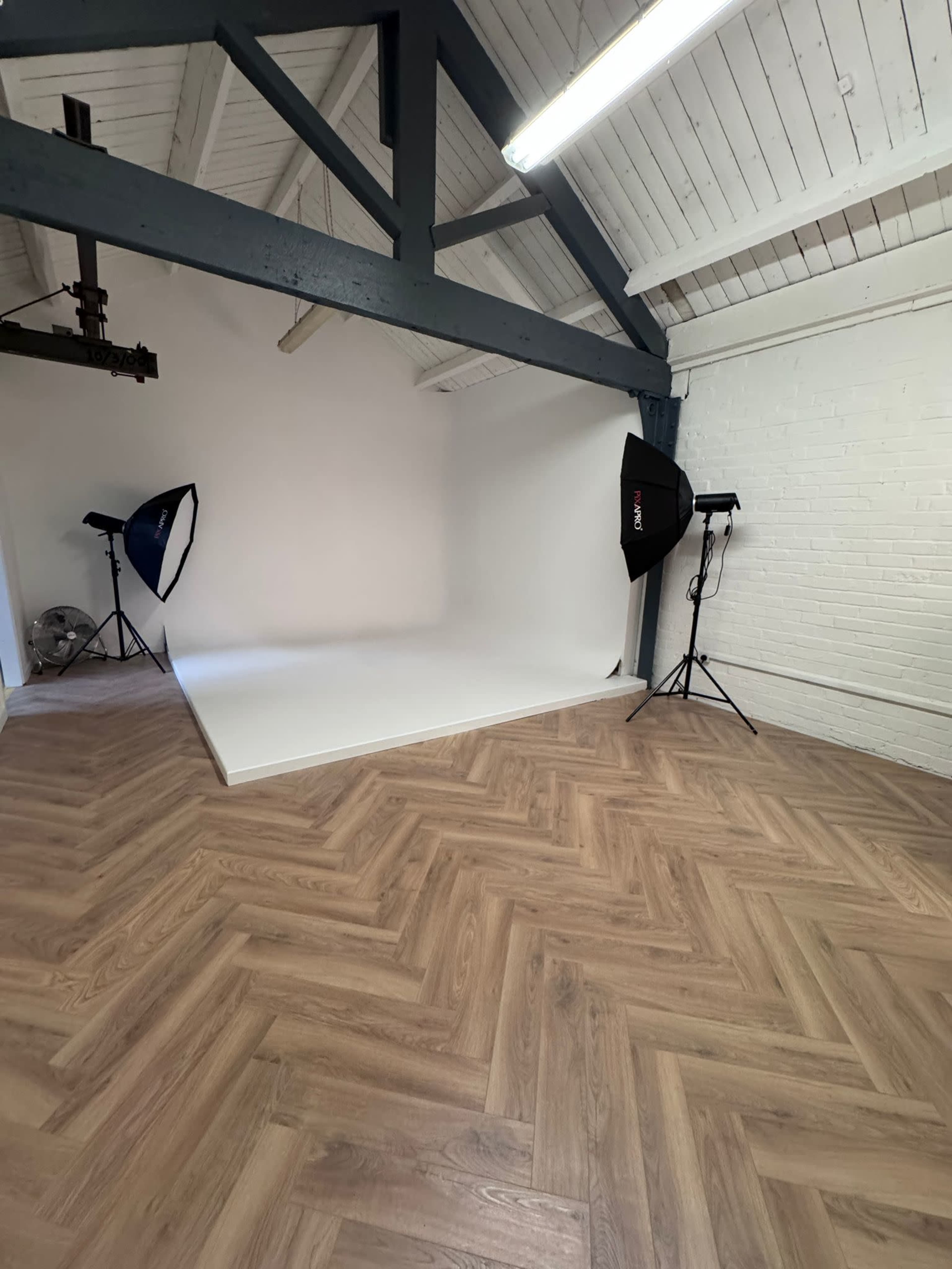 The image shows a photography studio with a large white backdrop, two light stands with softboxes, and wooden herringbone flooring.