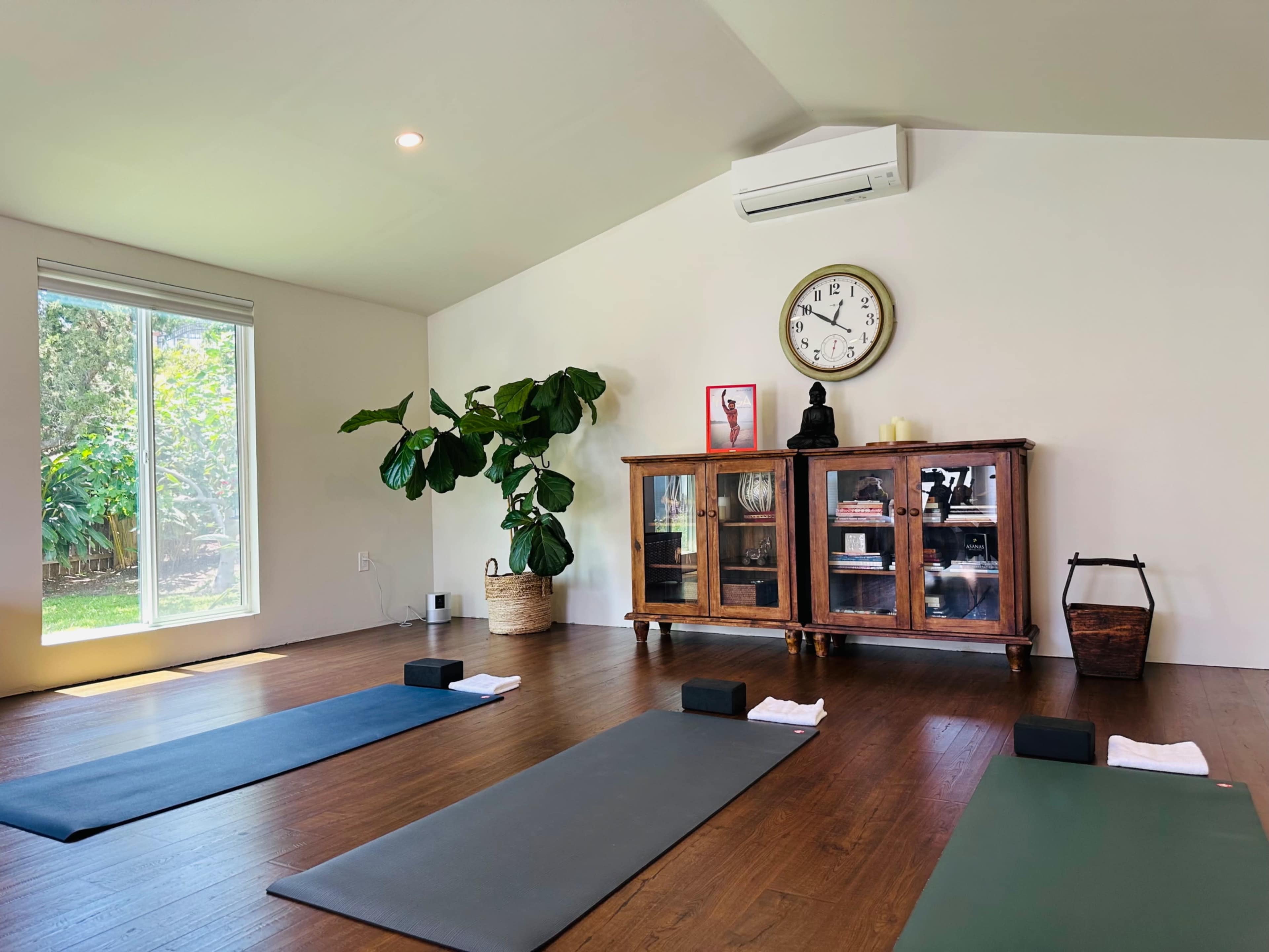 The image shows a calm yoga studio with two yoga mats laid out on a wooden floor, a clock on the wall, a Buddhist statue, and a cabinet filled with items, alongside a large indoor plant.