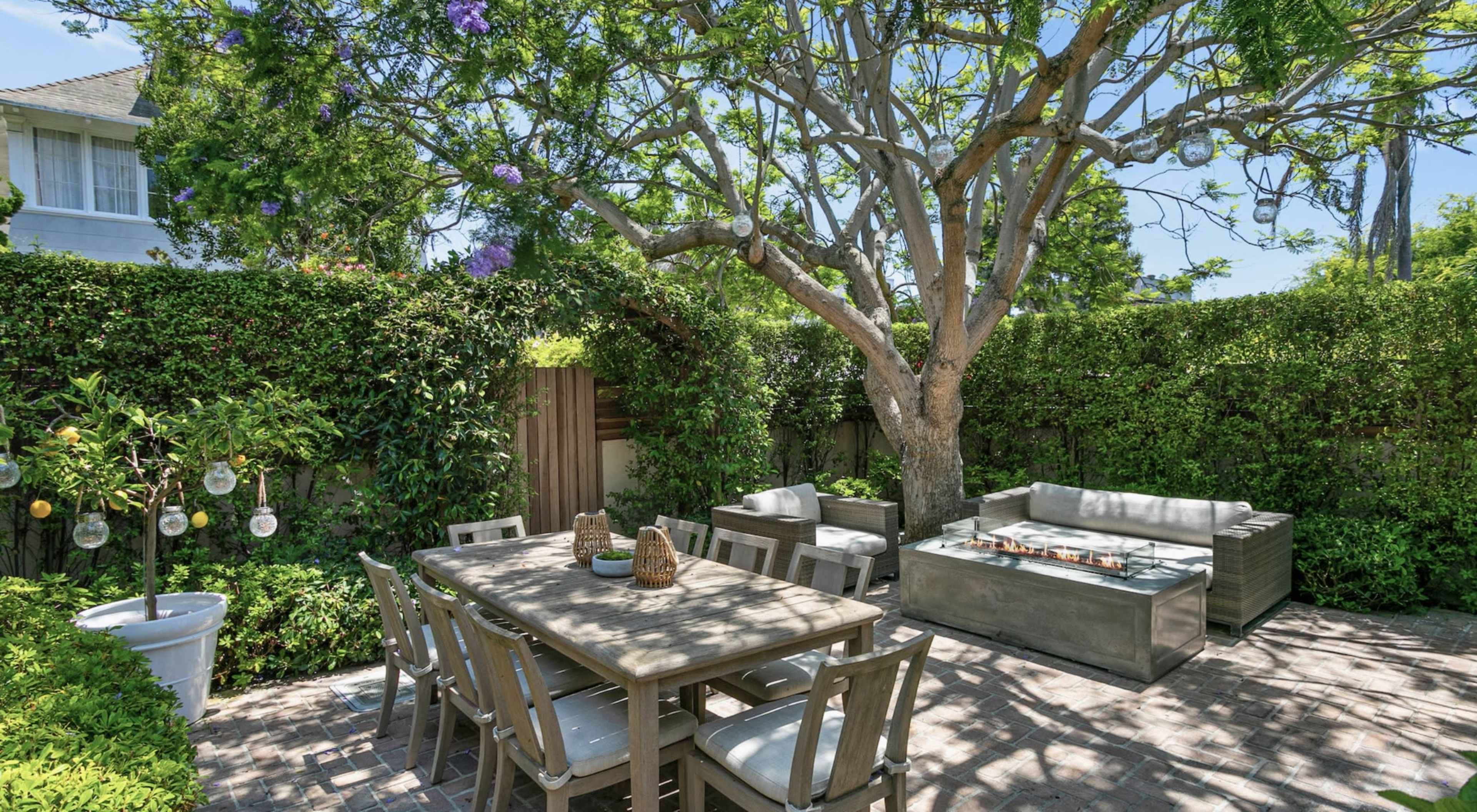 The image shows an outdoor patio with a dining table, seating area, and a large tree surrounded by hedges.