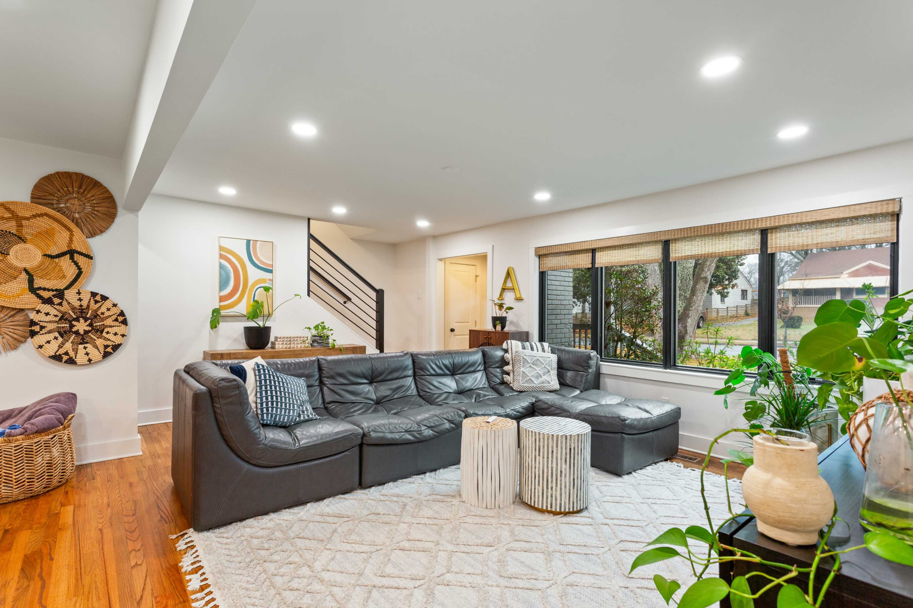The image shows a modern living room with a large sectional sofa, a woven rug, and large windows allowing natural light to enter.