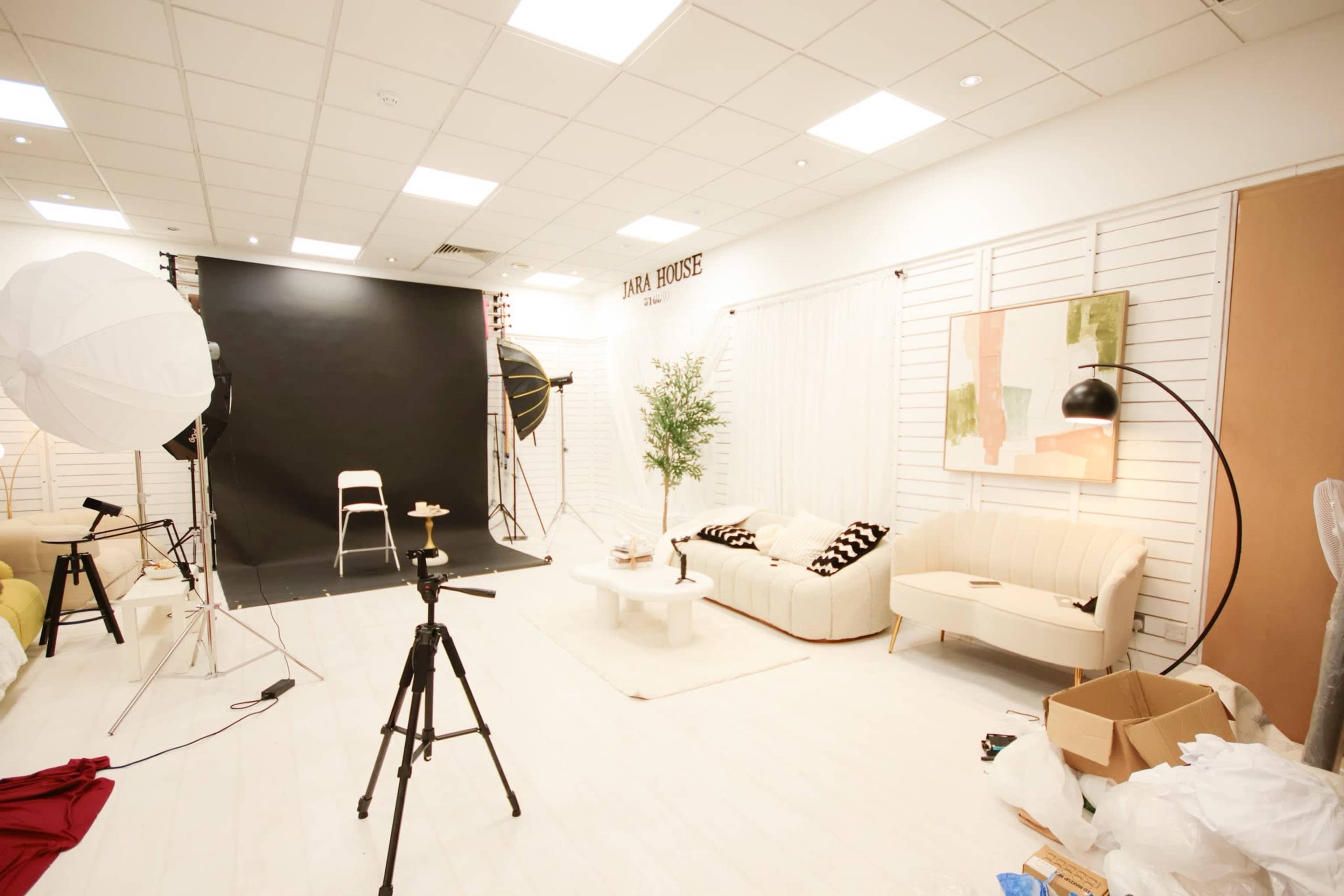 The image shows a photography studio with a backdrop, lighting equipment, a white sofa, and a decorative table.