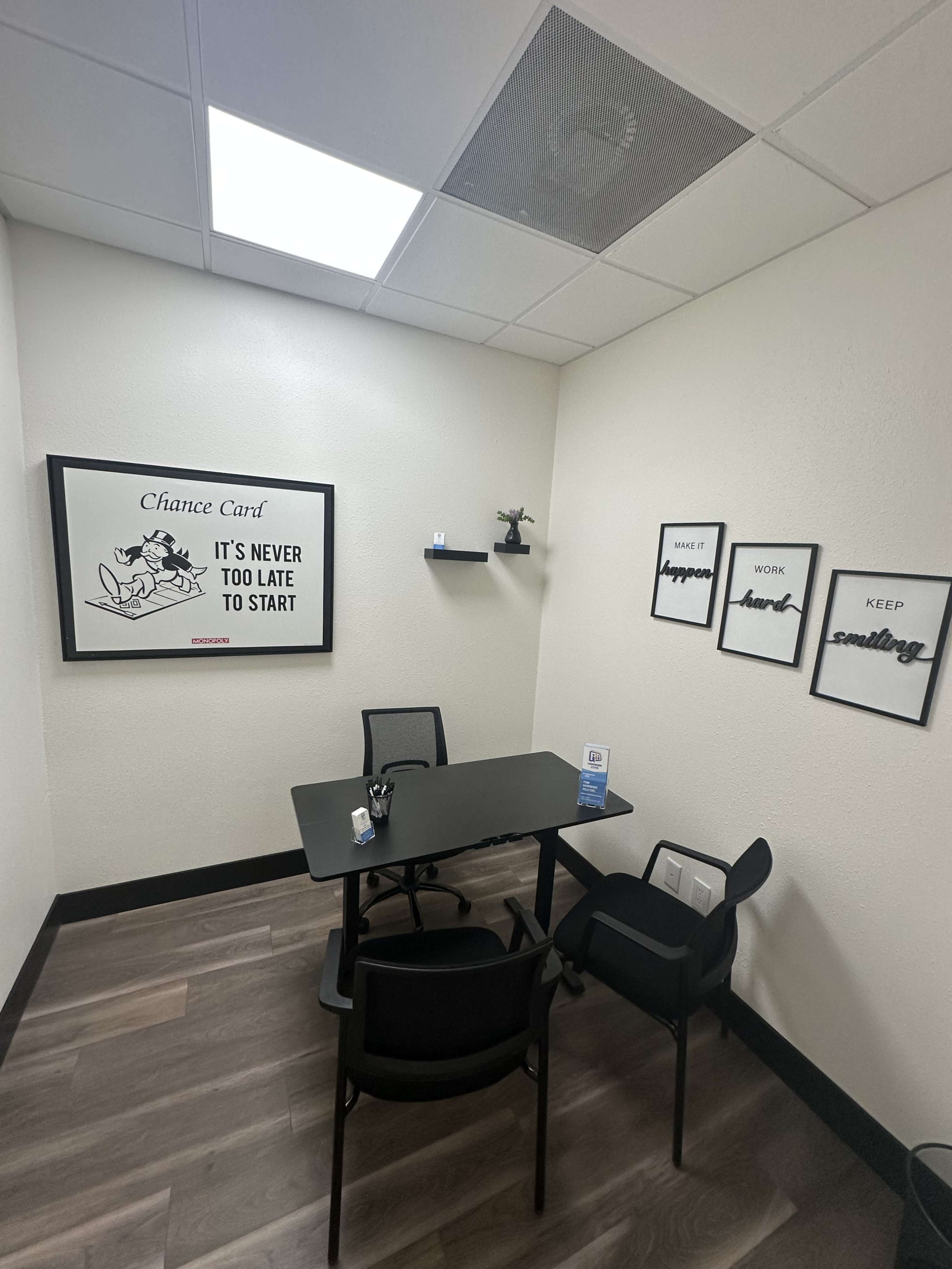 The image shows a small, lightly decorated office space with a black table and chairs, a motivational poster on the wall, and some framed quotes.