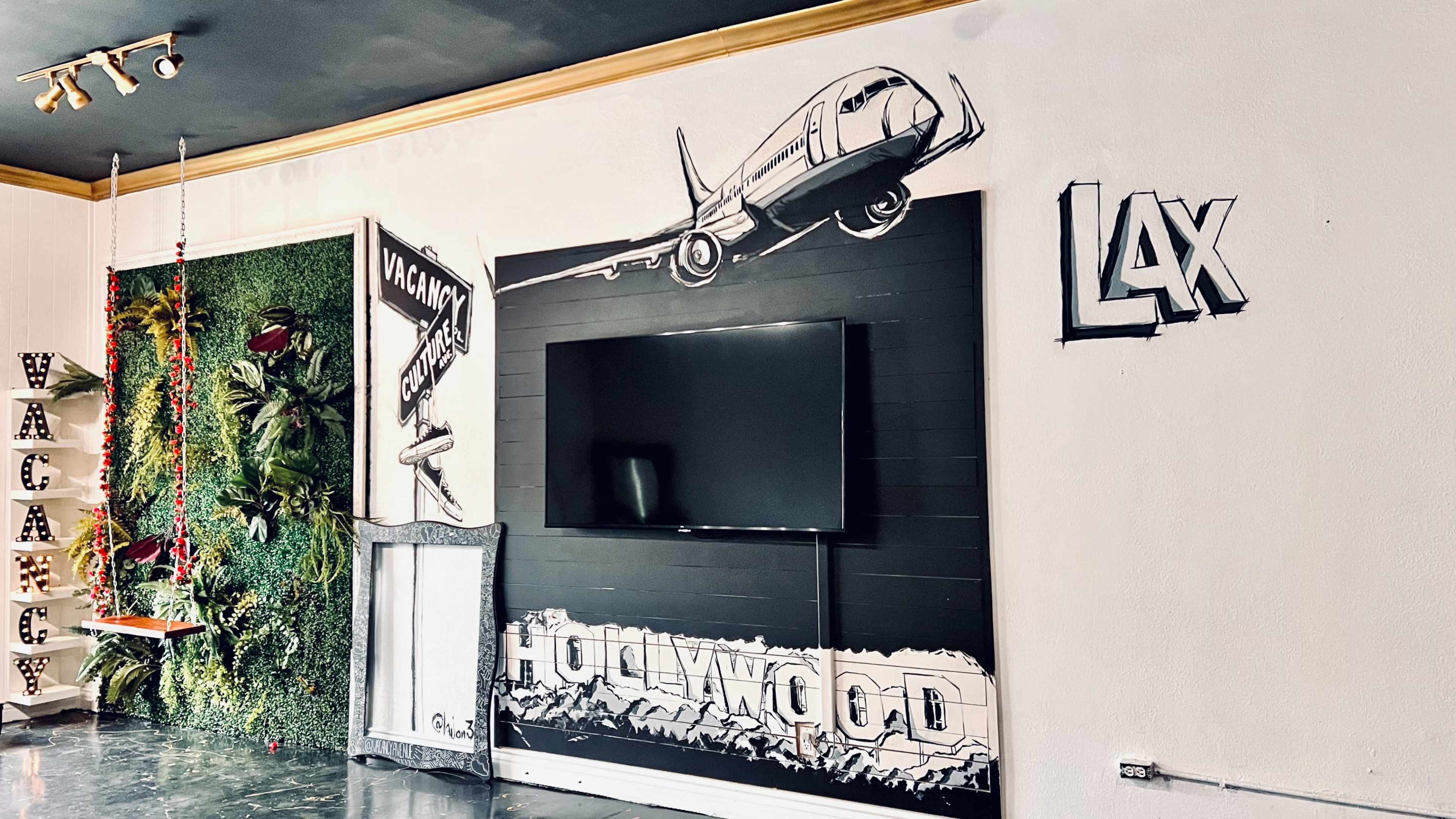 A wall decorated with a mural of an airplane and the words "HOLLYWOOD" and "LAX," alongside a TV and a green plant display.