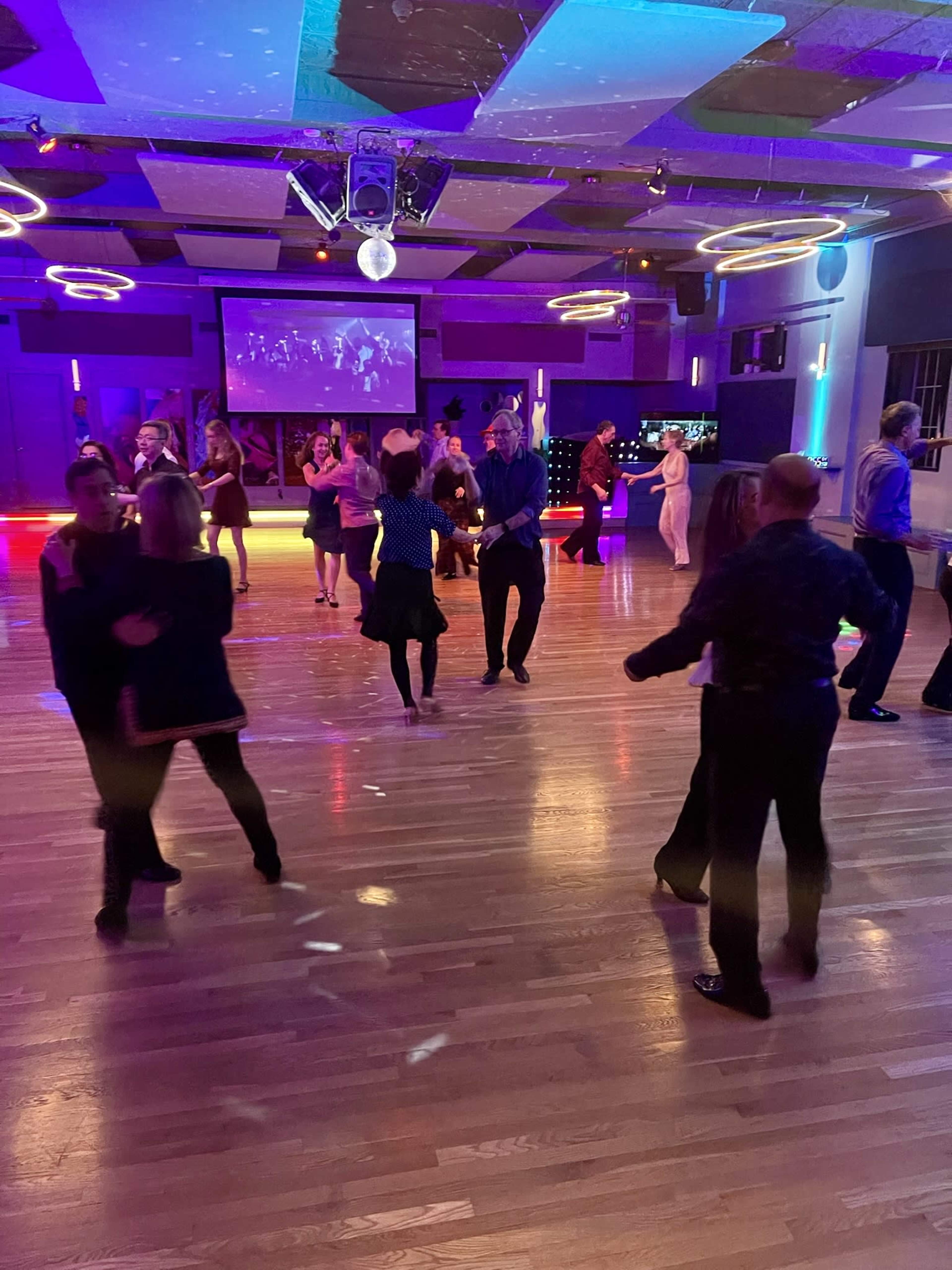 A group of people dance in a brightly lit hall with colorful lights and a large screen in the background.
