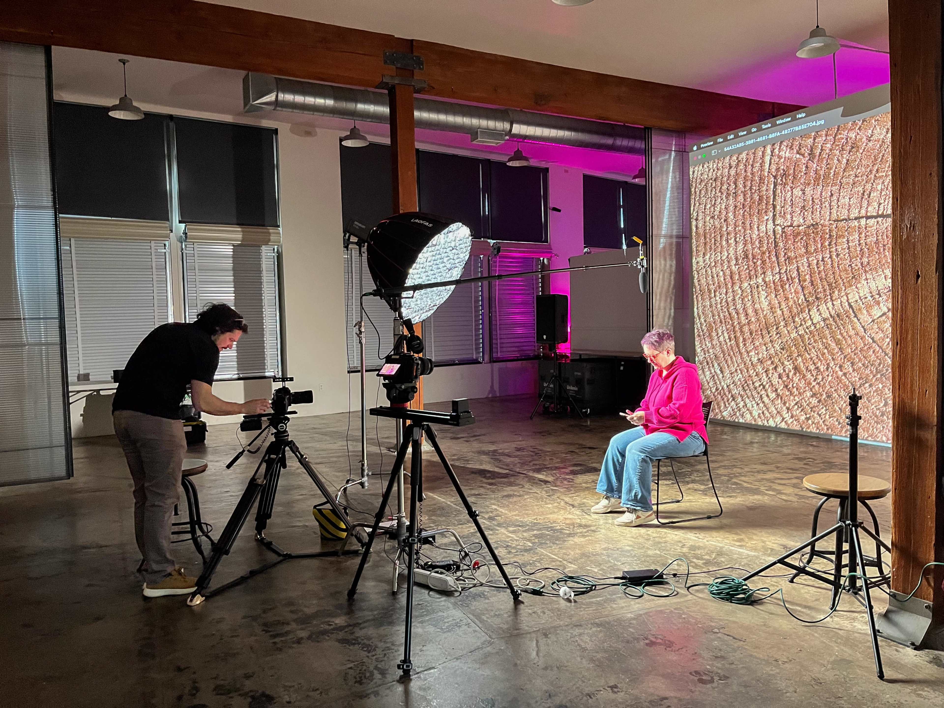 A person is sitting on a stool in a spacious studio, while another individual operates a camera to film them against a large backdrop displaying a textured surface.