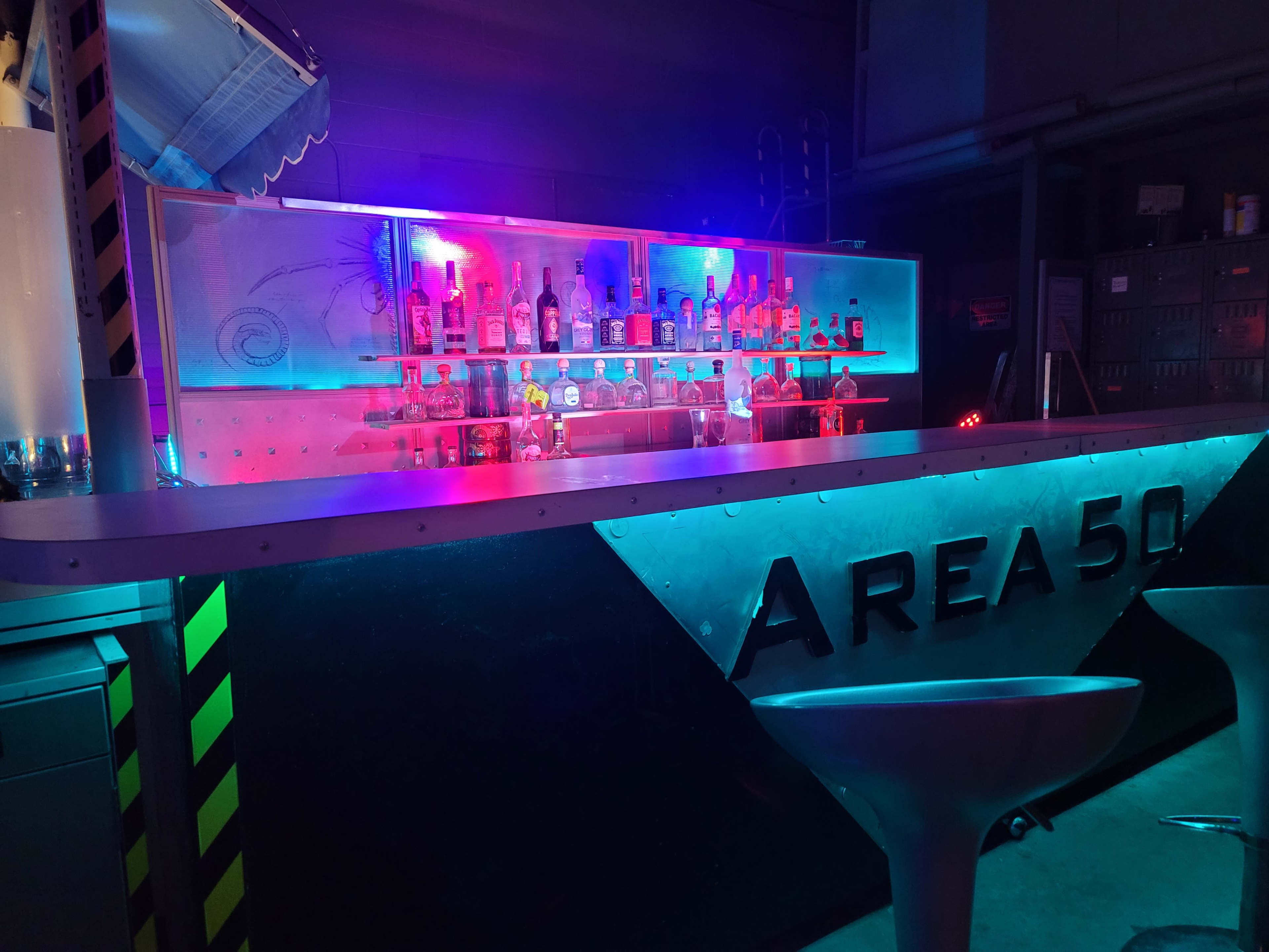 A bar labeled "AREA 50" with illuminated shelves displaying various bottles, set against a backdrop of blue and purple lighting.
