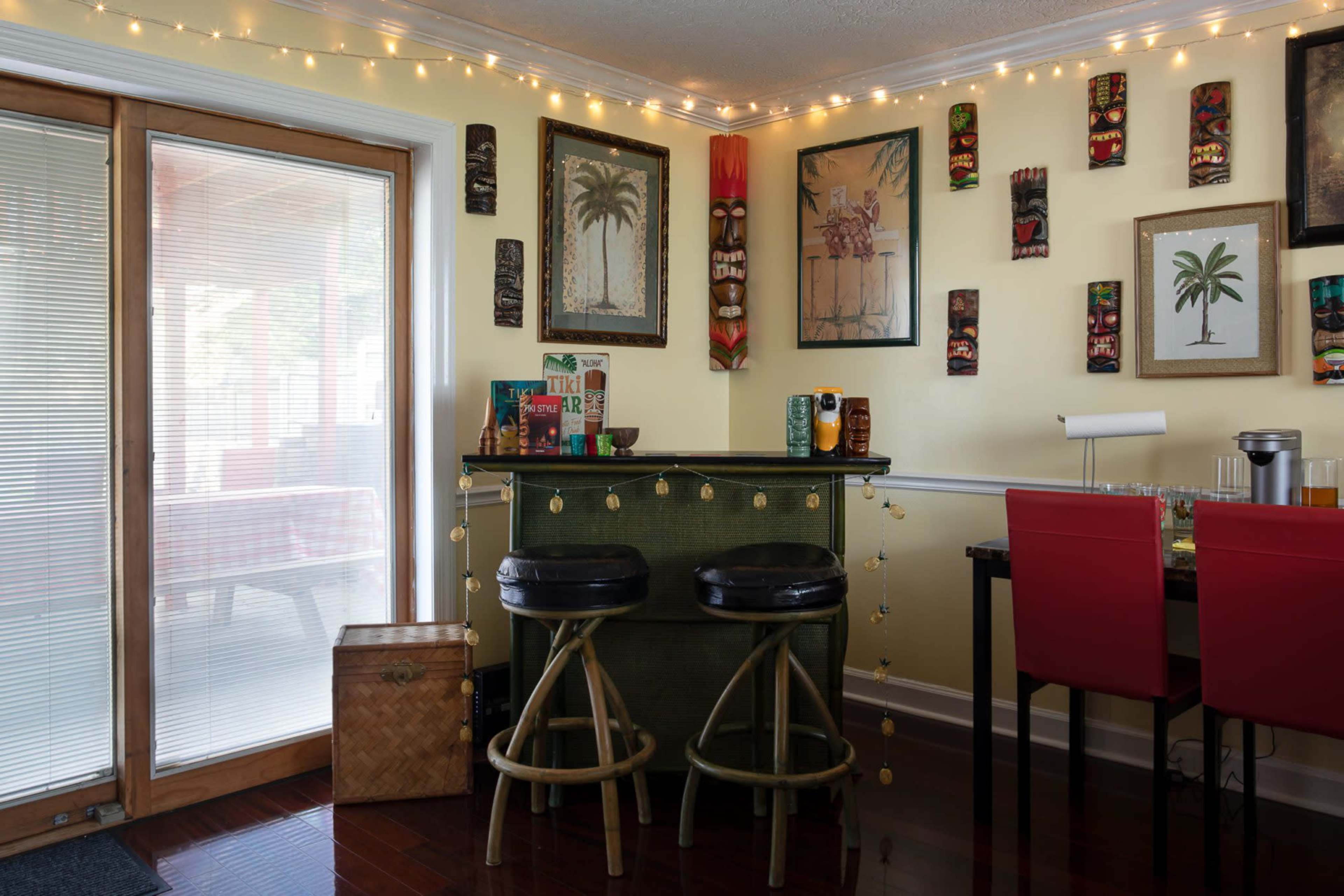 A small bar area with two stools is adorned with tropical-themed decorations, paintings, and string lights, beside a sliding glass door.