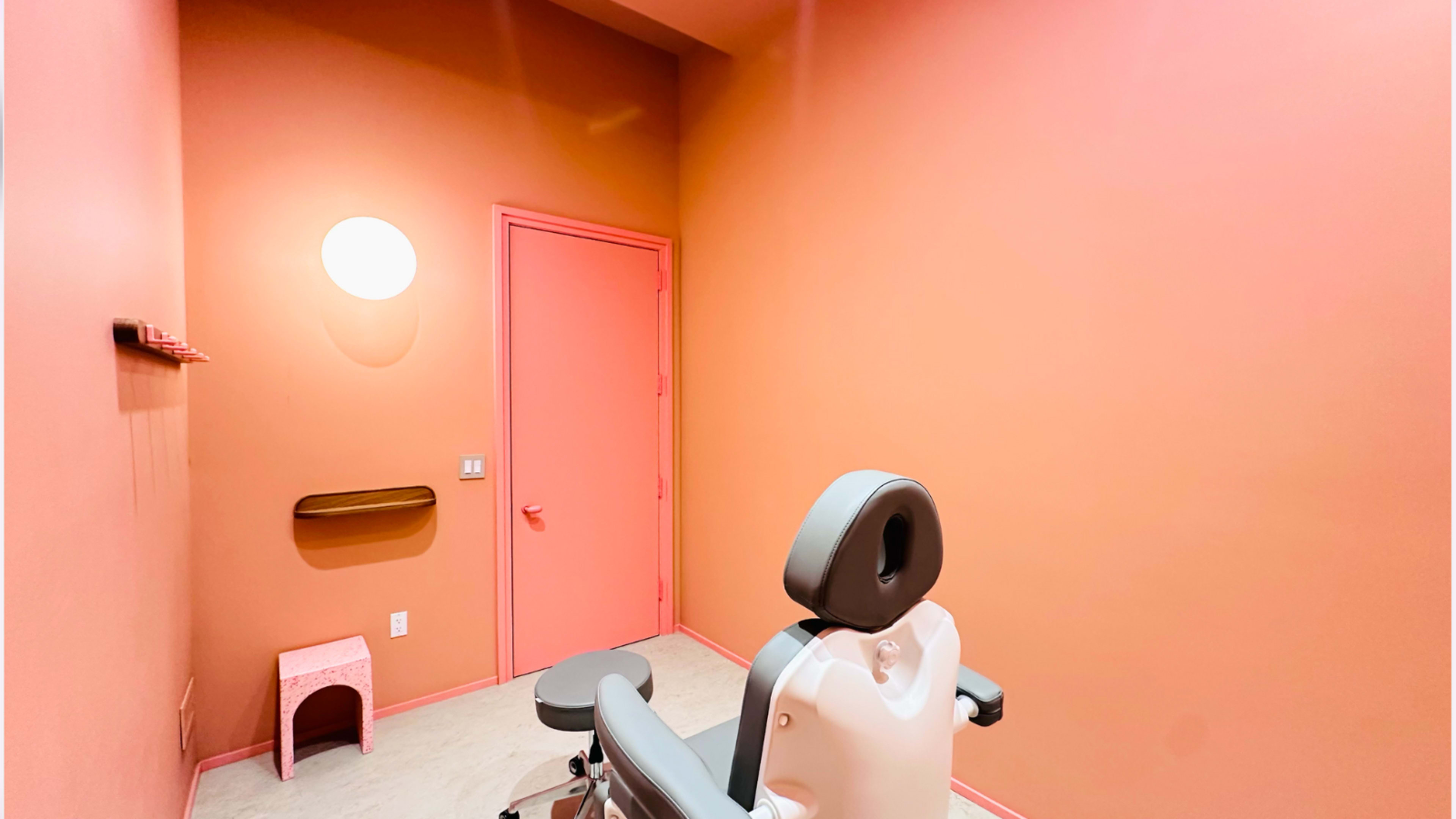 The image shows a small, minimalist treatment room with pink walls, a gray examination chair, and a closed door.