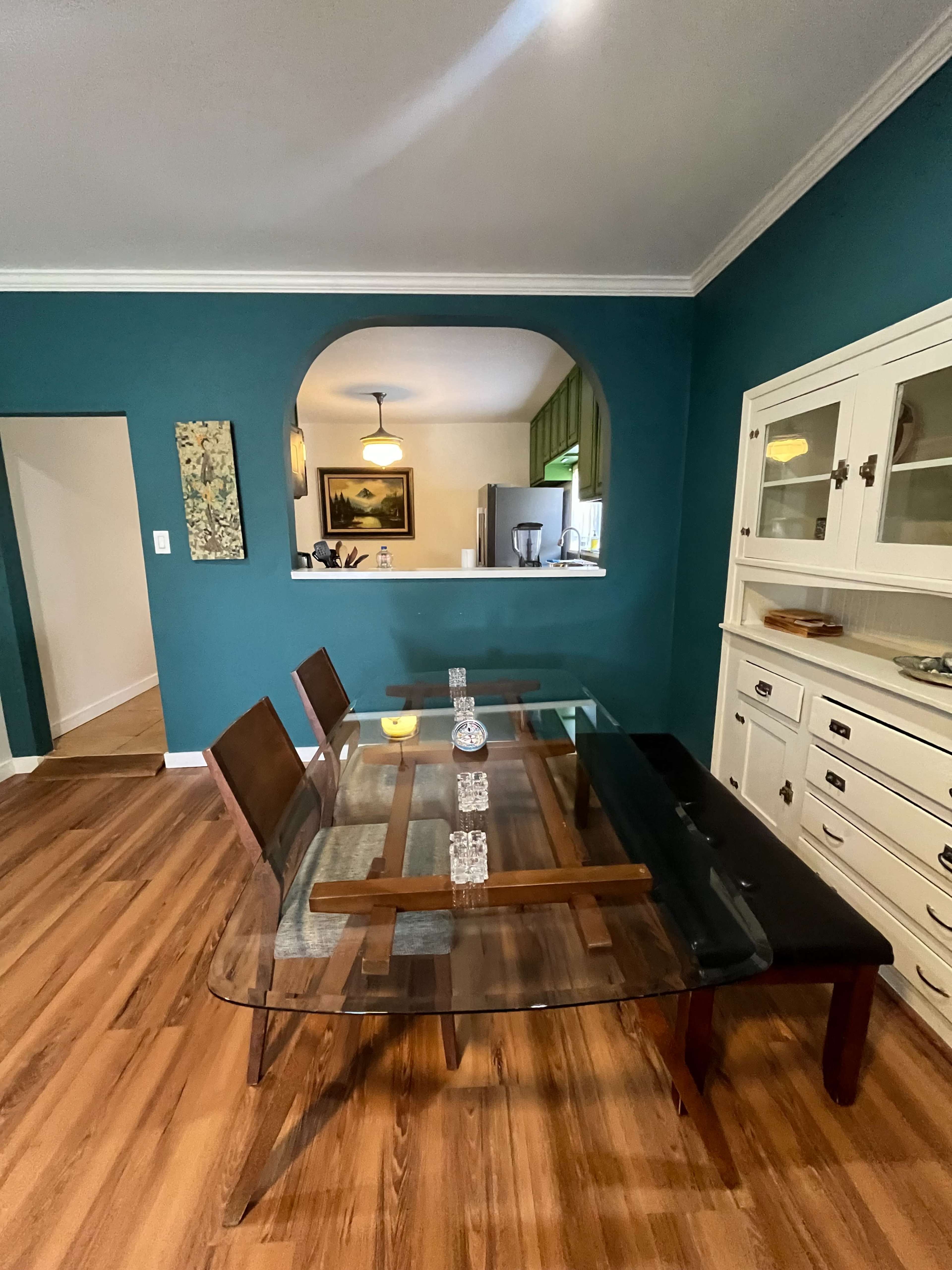 The image shows a dining area with a glass table and wooden chairs, accompanied by a sideboard against a teal-colored wall.