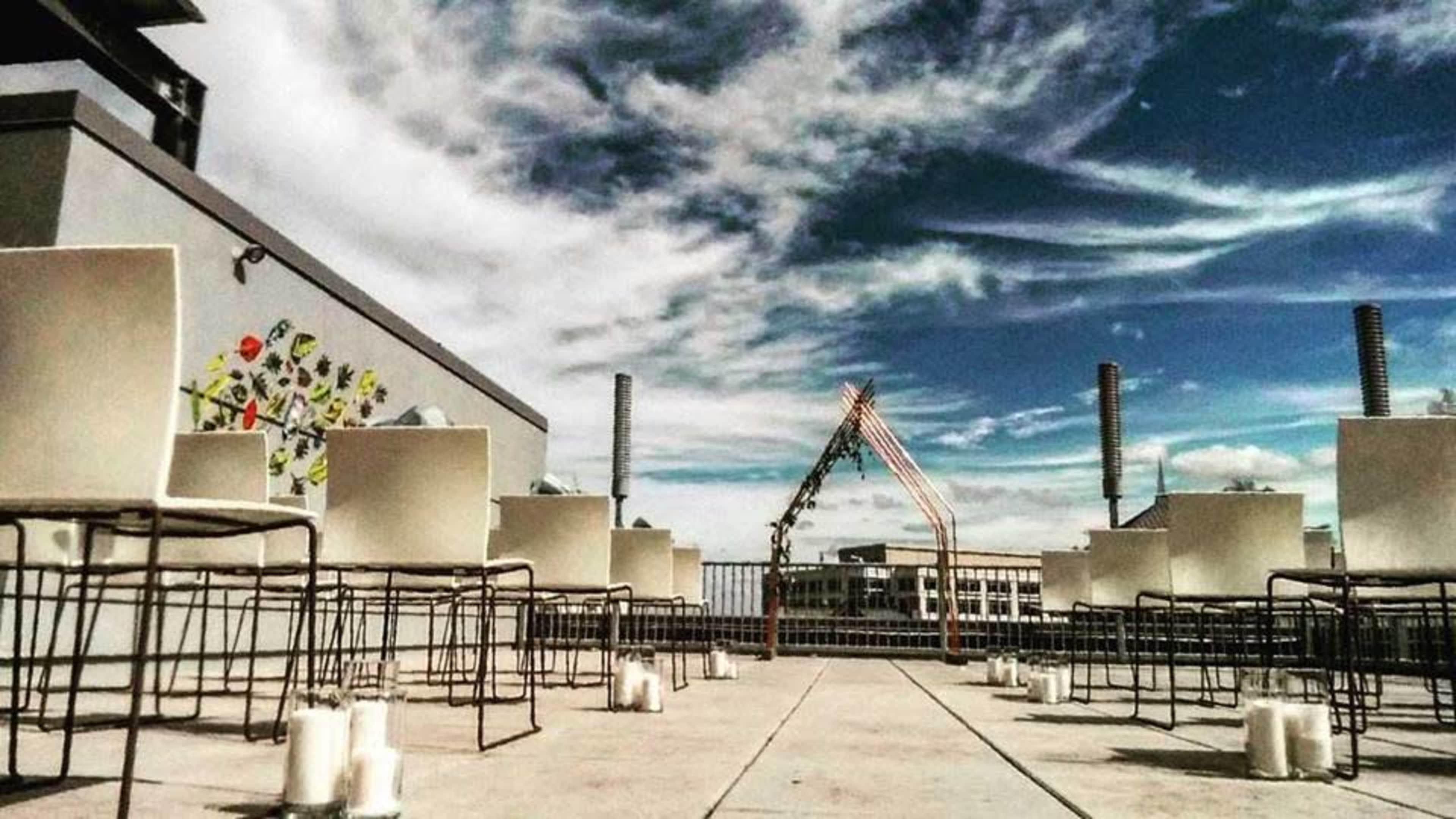 A rooftop venue is set up with white chairs arranged in rows, leading to an arch decorated with fabric, surrounded by candles on the ground.